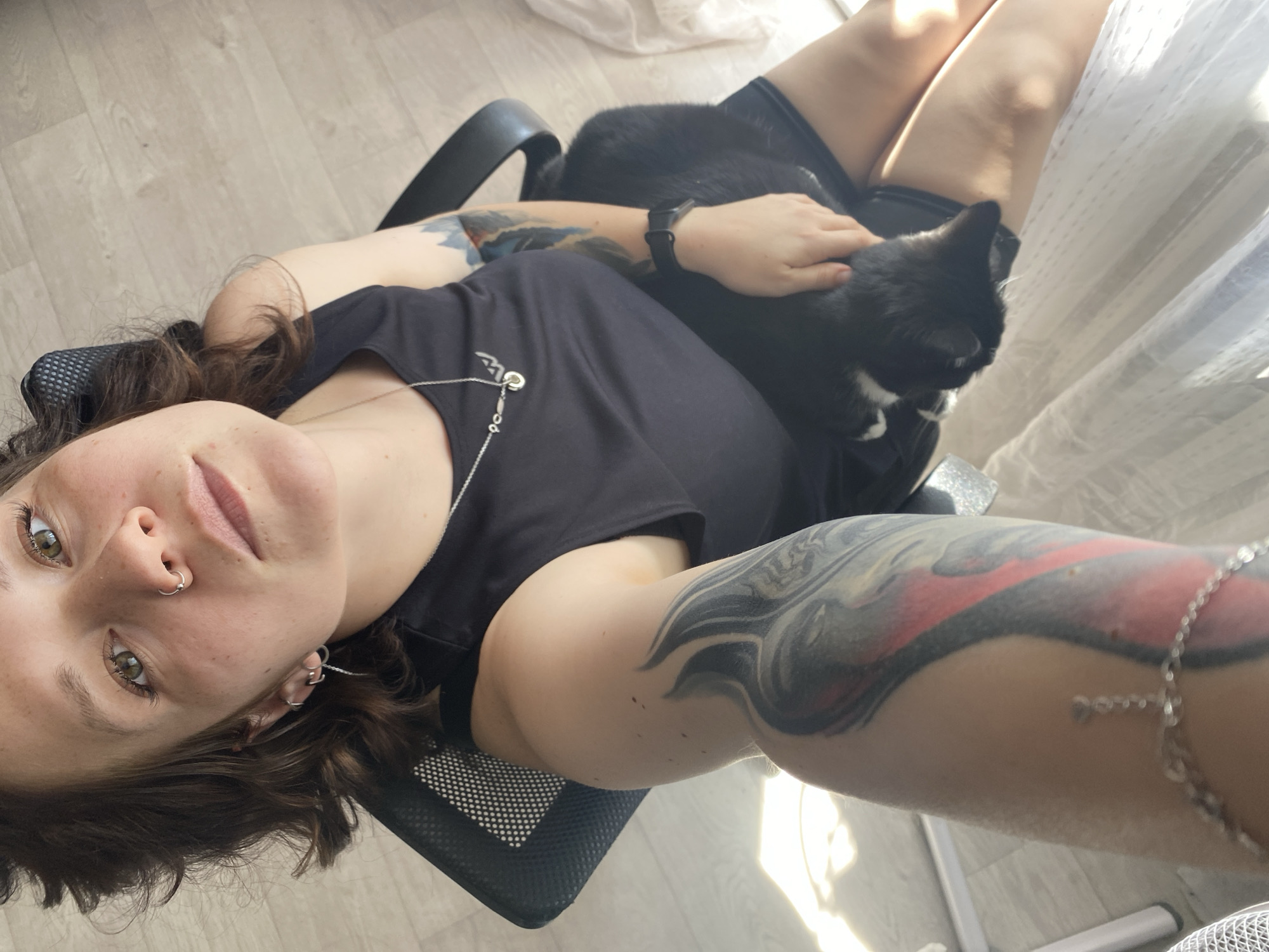 A selfie taken from a high angle shows a light-skinned woman sitting in an office chair. She is wearing a black tank top and has dark, curly hair. A black cat with white markings is sitting on her lap. She has tattoos on both arms and a silver necklace. The background shows a wooden floor, a curtained window, and part of the office chair. The location is Chaikovsky, Russia.