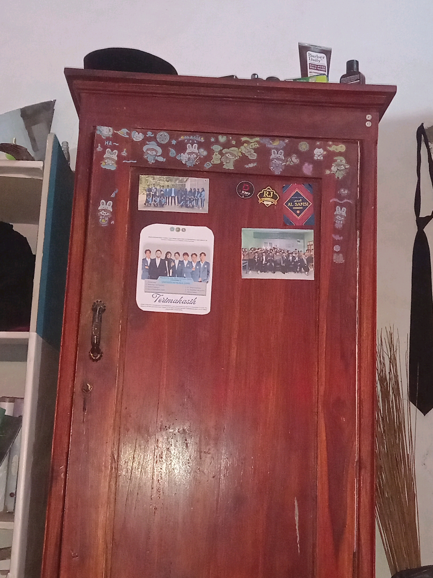 The image shows the top section of a dark reddish-brown wooden cabinet with several decorations and items placed on and affixed to its surface. A black hat is resting on the top edge of the cabinet. On the top surface, near the back, are two toiletries: a tube labeled "Barber Daily CAFFE-PORE REFINER" and a dark bottle with a black cap.
The front of the cabinet door is adorned with numerous stickers, primarily featuring cartoon characters, some of which appear to be rabbits and others in western-style attire. There are also three framed or printed items:
1. A small rectangular photograph above two stickers, depicting a group of people in uniforms standing outdoors.
2. A larger, rounded-top white print below the first photograph, showing a group of individuals in formal wear, with text at the bottom that reads "Terimakasih".
3. A square emblem with text that appears to be "RJ" and another with Arabic script and "AL SAMSI".
A decorative metal handle is attached to the left side of the cabinet door. To the right of the cabinet, a portion of a black tie is visible hanging down, and to the far right, a broom with natural bristles is leaning against the wall. The background is a plain white wall. The setting appears to be an indoor residential space, likely a bedroom or living area, in Randumerak, Indonesia.