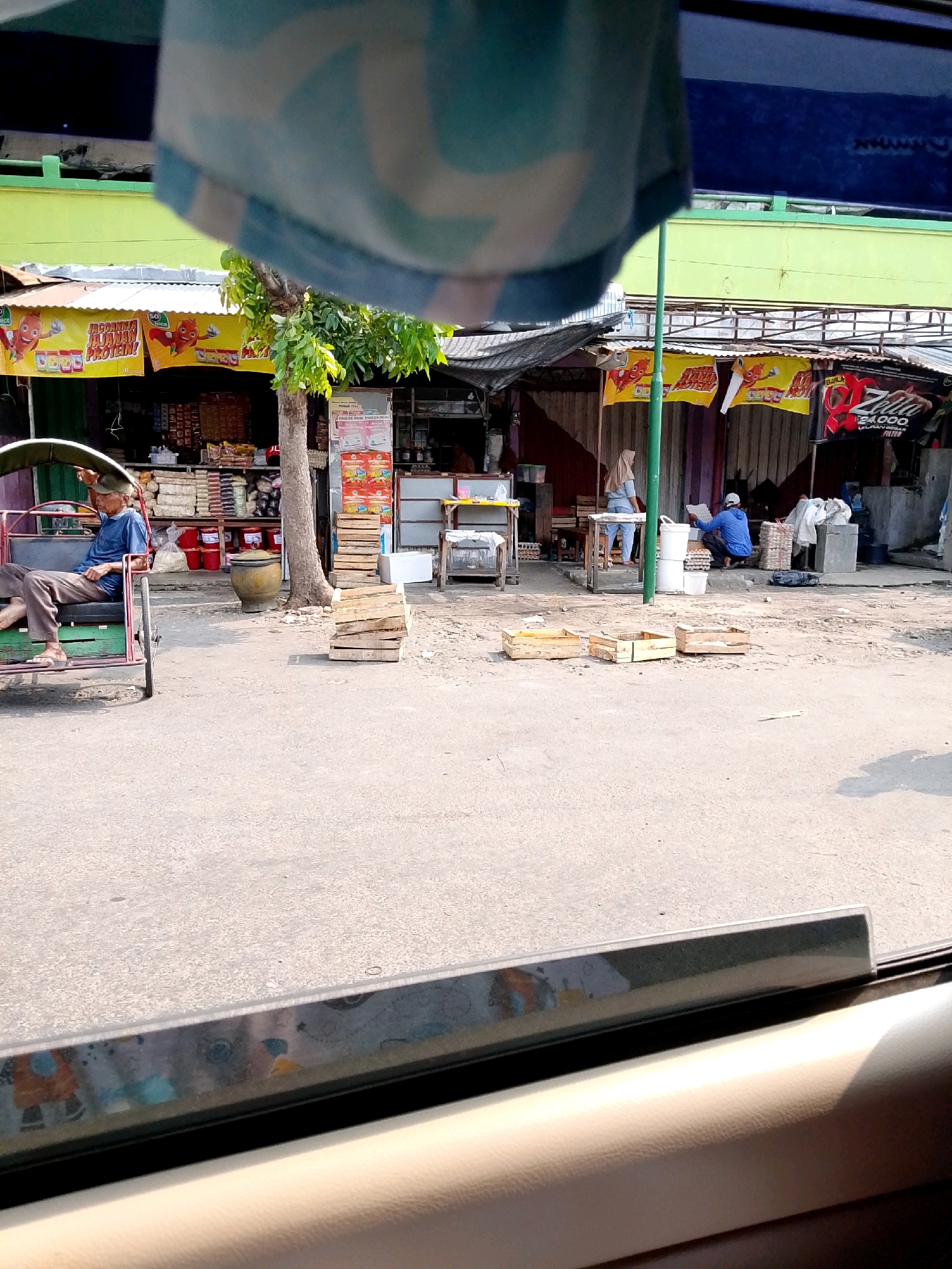 The image shows a street scene in Kediri City, Indonesia, with shops lining the road and several people present. In the foreground, a man sits on a pedicab. Behind him, a row of small shops with colorful banners display various goods. One banner advertises "Kacang Protein." Several people are seen interacting with the shops, including a woman in a hijab and a man wearing a blue shirt and a white cap. The environment appears to be a bustling market or commercial area. The overall impression is of a typical day in a vibrant Indonesian city.
