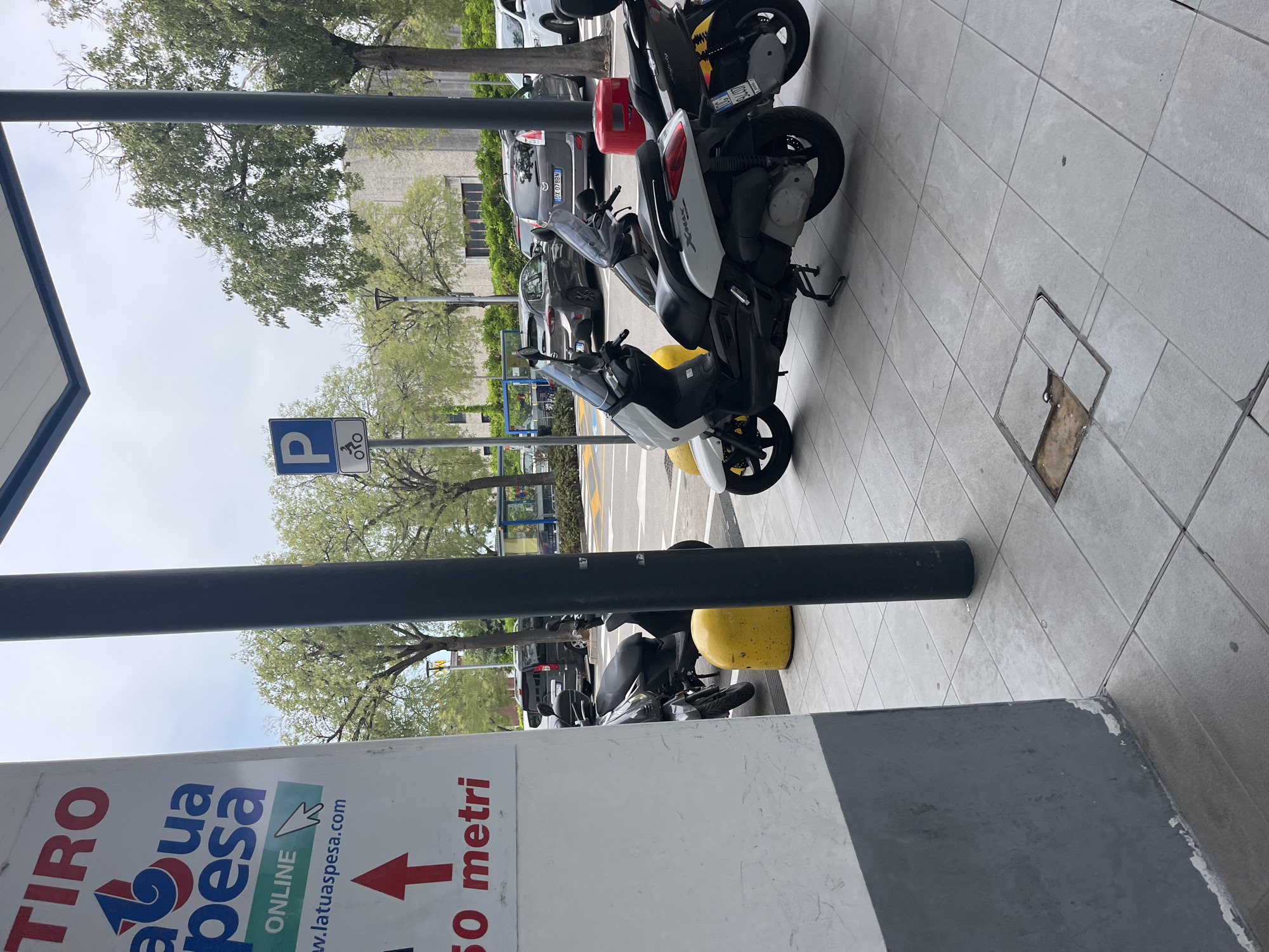 The image captures a scene in Castiglione delle Stiviere, Italy, featuring a paved area with several parked scooters and cars. In the foreground, a white and black scooter is prominently displayed, with another scooter partially visible to its right. To the left, a white pillar with red and blue lettering that reads "TIRO a tua spesa" and "ONLINE" is present, along with a red arrow pointing upwards and the text "50 metri". Further back, a yellow cylindrical object, possibly a bollard, is situated near a dark-colored scooter. A blue and white parking sign with a "P" and a bicycle symbol is visible in the background. Several vehicles, including a dark SUV, a grey hatchback, and a white van, are parked in the distance. The environment appears to be an outdoor public space, possibly a parking lot or commercial area, with trees and buildings visible behind the parked vehicles. The sky is overcast, suggesting a cloudy day.