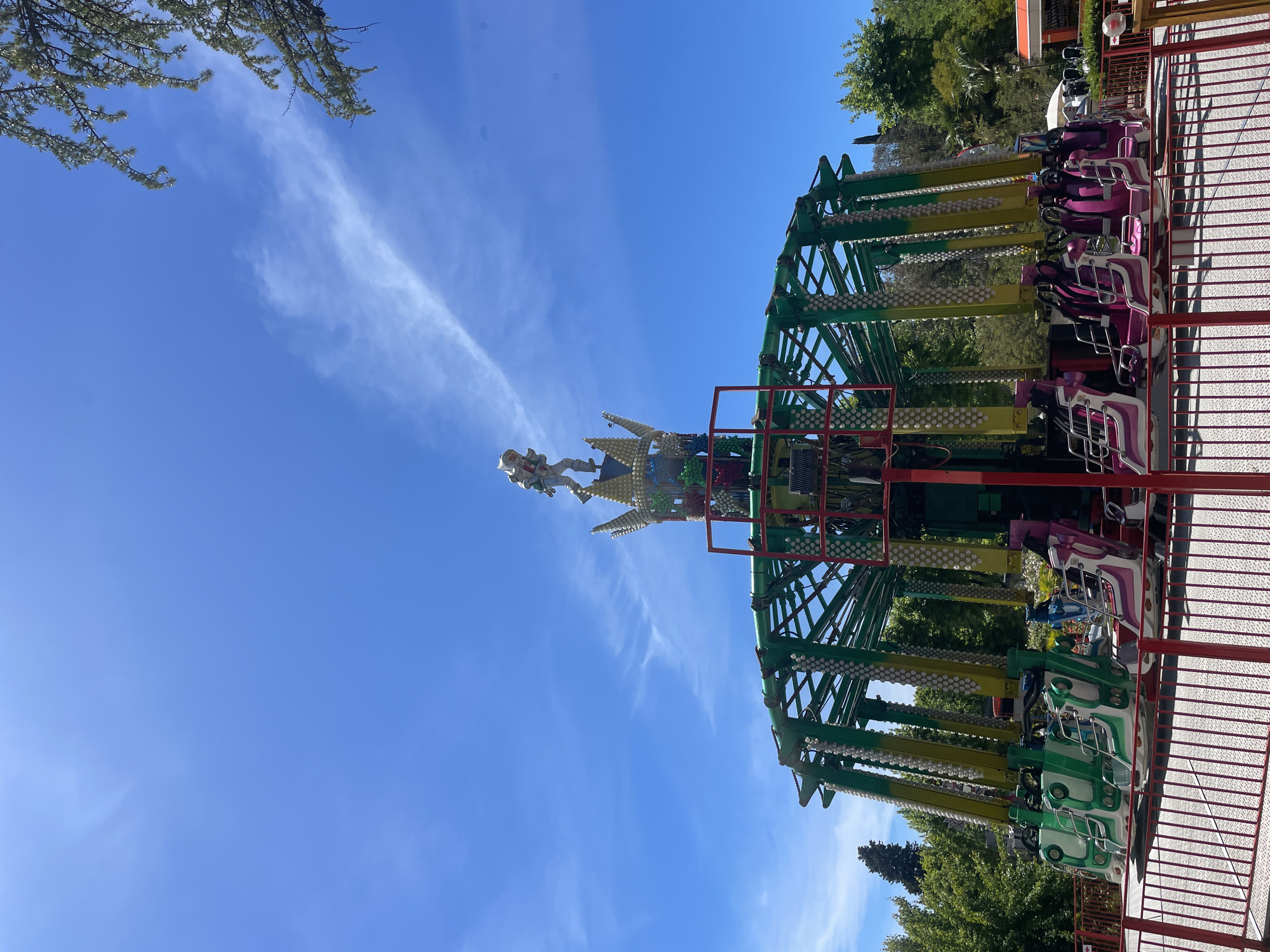 The image, correctly oriented, displays a large amusement park ride under a bright blue sky with wispy white clouds. The ride is a swing-style attraction, featuring a prominent green and yellow metal structure adorned with numerous small, white spherical lights or decorations. It has multiple gondola-like seats, which are currently empty. Some seats are distinctly pink and purple, while others are green and white.
At the apex of the ride's central column, a decorative element resembling a crown or spire is visible, featuring several figures. One figure, dressed in white and appearing to be an astronaut or knight, is clearly distinguishable. The ride is positioned outdoors, with dense green trees and foliage visible in the background, suggesting a park setting. In the foreground on the right, a red metal fence or railing provides a boundary for the attraction.
The scene depicts a sunny day, likely mid-day or early afternoon, given the strong illumination and clear sky. The ride appears to be stationary, with no people actively riding it or in the immediate vicinity, implying it is either closed, awaiting operation, or between cycles. The overall atmosphere suggests a leisure environment at an amusement park in Sochi, Russia. There is no visible text in the image.