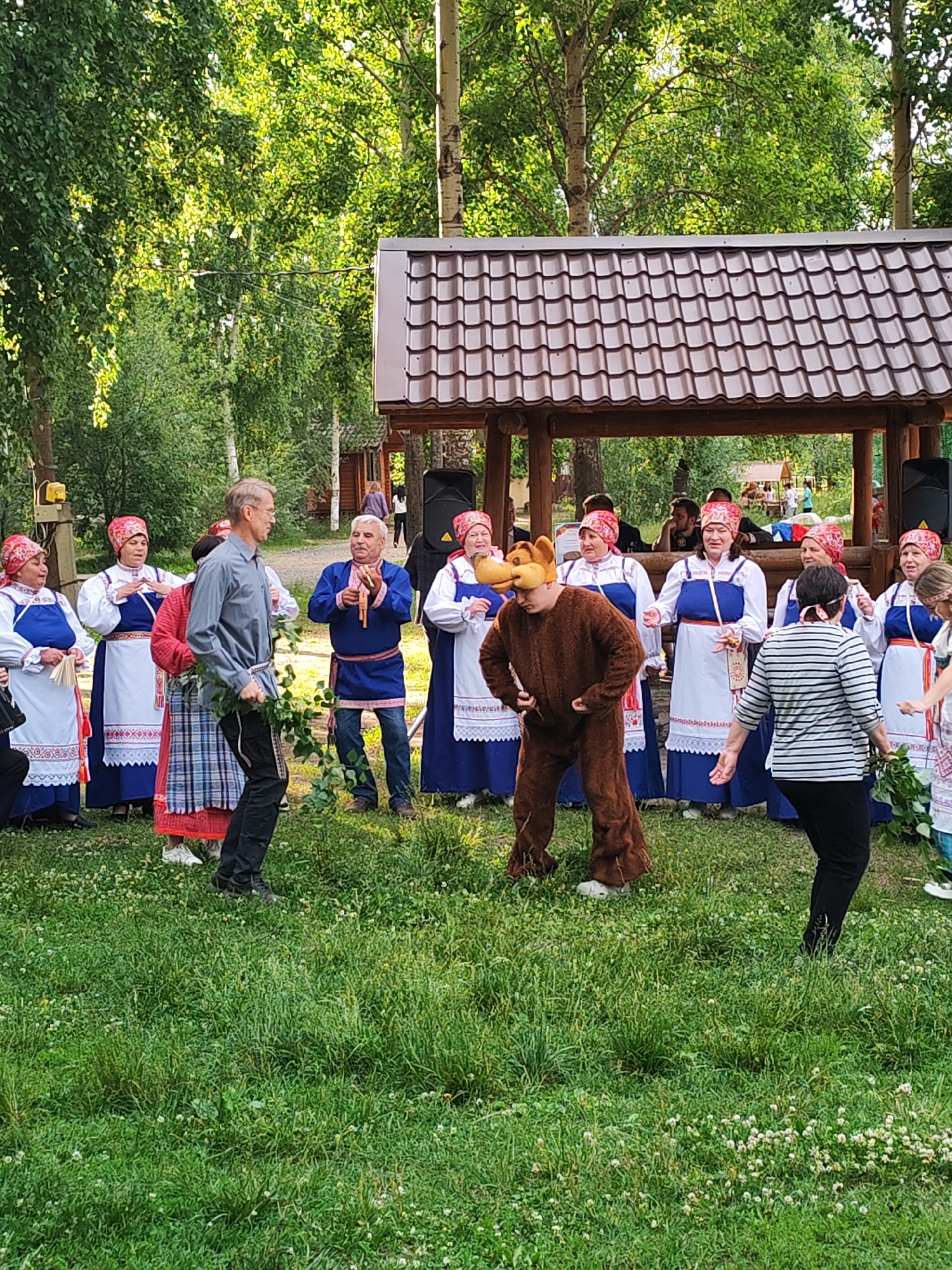 In a grassy outdoor area in Chaikovsky, Russia, a group of people are gathered, seemingly for a festive event. Several women in traditional attire, characterized by blue dresses, white aprons with red embroidery, and red headscarves, stand in a semi-circle. One woman is holding a bottle. A man in a grey shirt and black pants, holding a bunch of greenery, is walking towards the group. To his left, a man in a blue tunic and dark pants plays a wooden flute-like instrument. In the center, a person in a furry brown bear costume with a distinctive headpiece is standing, with their hands on their hips. Another woman in a black and white striped shirt and black pants is dancing, facing the bear. The background features a wooden pavilion with a brown tiled roof, and lush green trees, suggesting a sunny day, possibly late afternoon given the warm light. The overall mood appears to be joyful and celebratory, indicative of a cultural performance or folk festival.