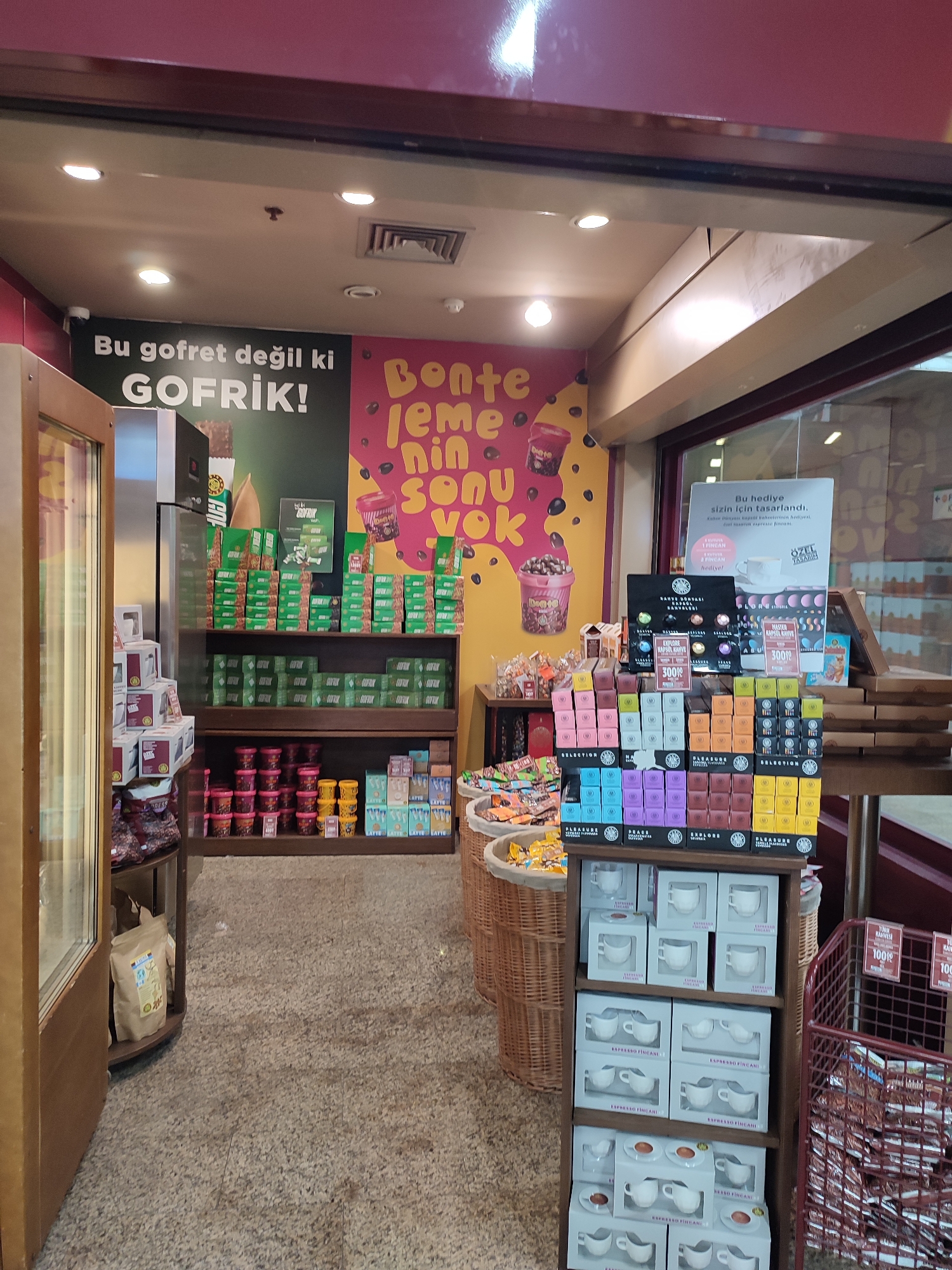 The image captures the interior of a cafe or shop, likely a chain store, in the Beyoğlu district of Istanbul, Turkey. The scene is an indoor retail environment with a focus on coffee-related products. In the foreground and mid-ground, shelves are filled with various packaged goods, primarily coffee pods in colorful boxes, small tubs of what appear to be coffee or confectionery, and bagged items. A prominent display features sets of small white cups and saucers, arranged in boxes. To the left, a large glass-fronted cabinet or display case is visible, with some bagged items inside. A large promotional poster with Turkish text, featuring the word "GOFRİK!", is displayed on the wall above the shelves. The overall impression is one of a well-stocked retail space designed to sell coffee and related accessories.