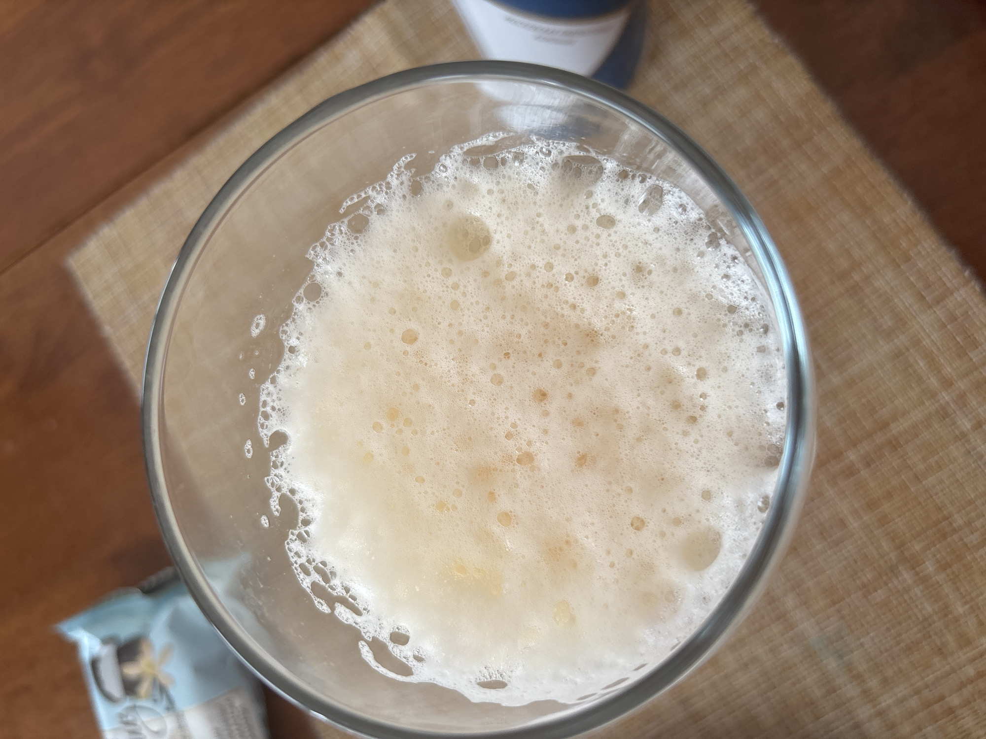 A close-up, overhead shot shows a glass full of a frothy, pale yellow beverage, possibly beer or a soft drink, with a thick layer of foam on top. The glass sits on a woven placemat, which is on a wooden surface. In the background, out of focus, are what appear to be a package of cookies or biscuits and a blue and white bottle. The lighting suggests an indoor setting, likely a home or cafe. There are no people or animals in the image. The scene is devoid of any specific markers to indicate a location in Sochi, Russia.