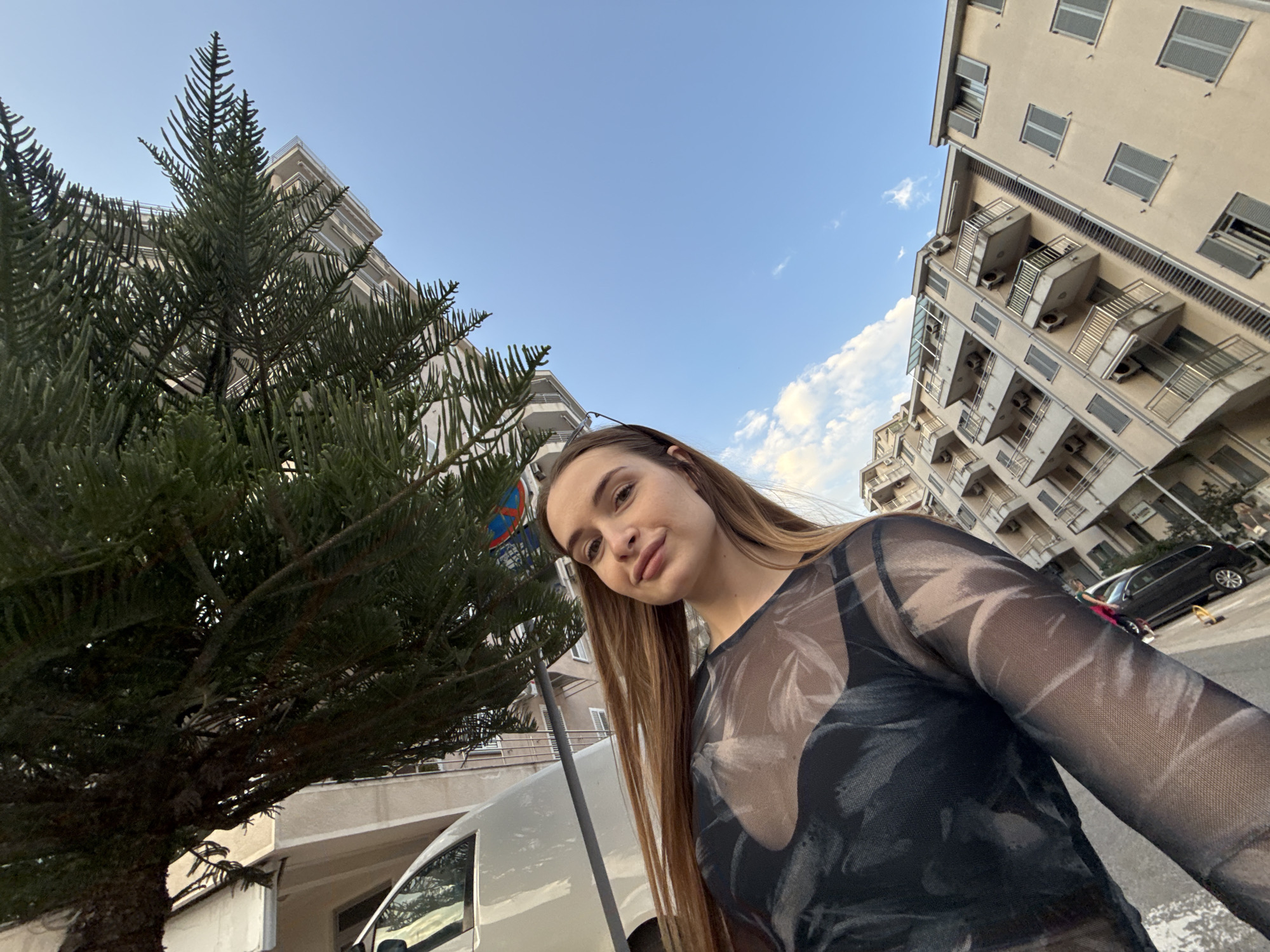A low-angle selfie depicts a young woman in an urban setting. She wears a black, sheer, long-sleeved top. Her long, light brown hair hangs down to her chest. She looks directly at the camera with a slight smile.
To her left is a large green tree with dense foliage. Behind the tree and to the left of the woman is a light-colored building. To her right, there is a multi-story building with balconies, a black car, and a van. The sky is blue with some clouds.
The location is likely in Budva, Montenegro, given the architecture of the buildings and the vegetation.
