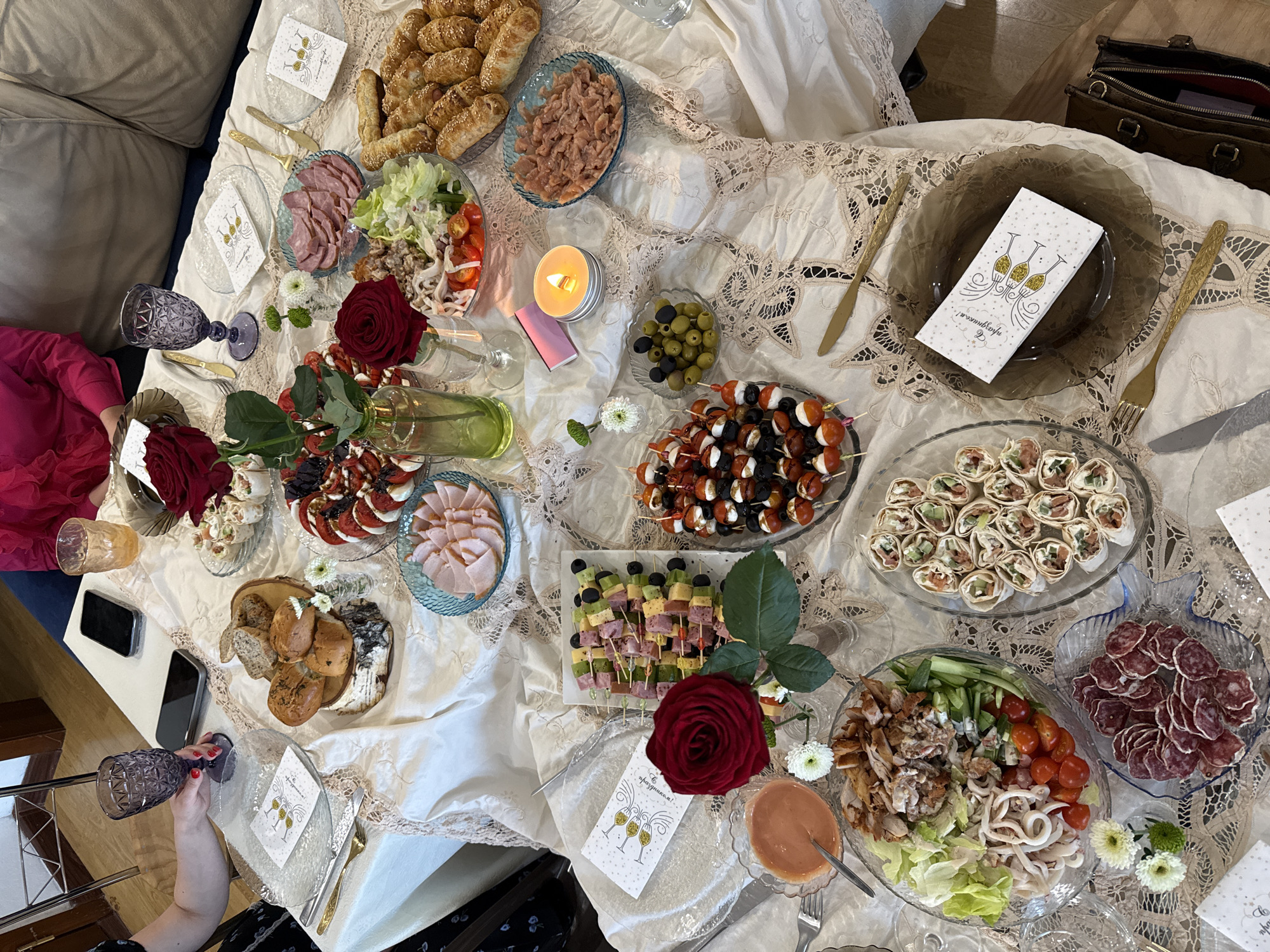 The image captures a festive table spread, likely indoors, featuring a variety of dishes indicative of a celebratory occasion. The table is covered with a textured, possibly embroidered, white tablecloth. Numerous plates hold different types of food: small pastries, cured meats, salads, skewers with olives and tomatoes, ham slices, tortilla rolls, and bread. Decorative elements include roses and small white flowers. Glasses and cutlery are set around the table. Napkins or cards with a champagne glass design are present. A lit candle adds to the ambiance. At least one person is visible, holding a purple-tinted glass. In the background, part of a sofa and a handbag can be seen. There is no indication that the location is in the city of Krasnoyarsk, Russia.