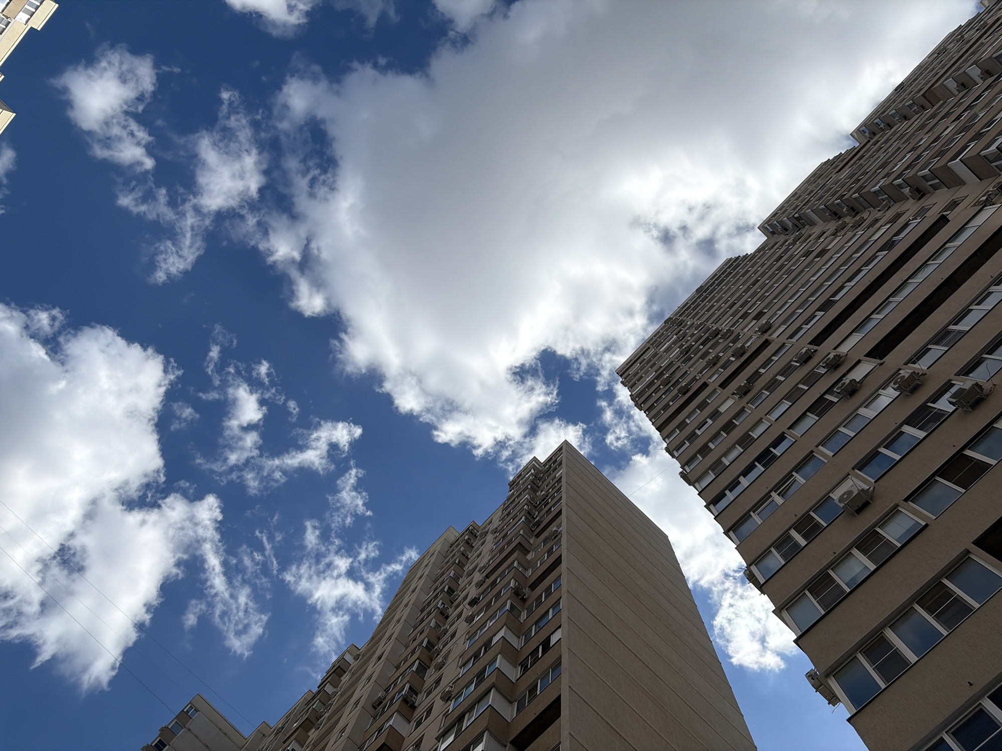 This image, captured in Krasnodar, Russia, depicts a low-angle perspective looking upwards between two tall, multi-story residential buildings against a partly cloudy blue sky. The buildings are primarily beige or light brown, characterized by numerous windows and externally mounted air conditioning units visible on their facades. The building on the left features a distinctive stepped or zigzag architectural pattern along its visible edge. Three thin, dark utility lines traverse the upper left quadrant of the sky. The sky itself is a vibrant blue, populated by scattered white cumulus clouds, with a larger, denser cloud formation occupying the upper right portion of the frame. The scene is devoid of people, vehicles, or specific ground-level elements, emphasizing the verticality of the structures and the expansive sky above. Illumination suggests clear daytime conditions.