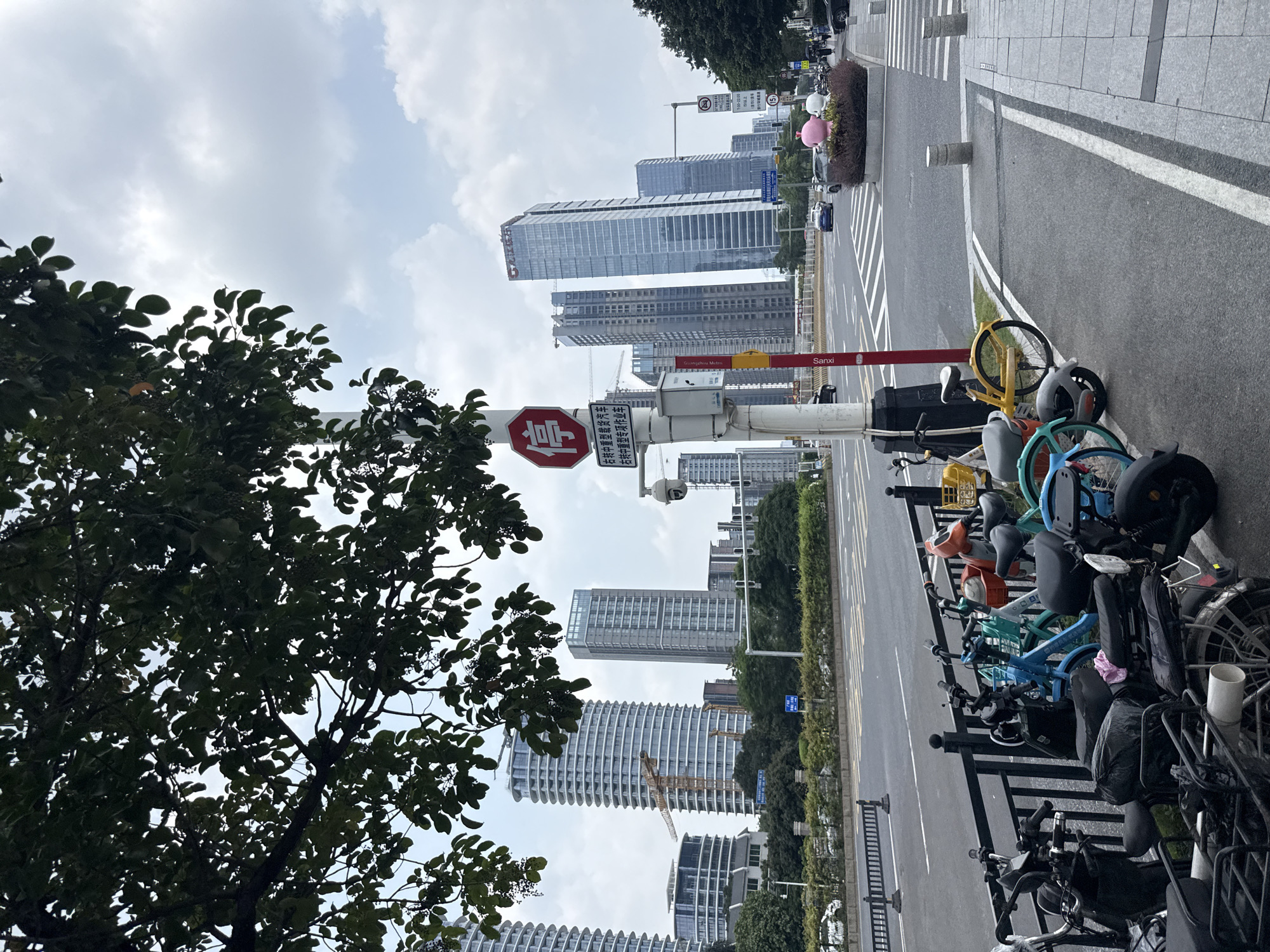 This media file captures a vibrant urban street scene in Tianhe District, China, on what appears to be an overcast or partly cloudy daytime. The image is composed of a mix of natural elements and man-made structures, illustrating a bustling city environment.
In the foreground, lush green trees with dark foliage occupy the left side, partially obscuring the view of the sky. On the right, a row of shared bicycles, varying in color (yellow, blue, orange, black, grey), are neatly parked along a black metal railing beside the sidewalk. The bikes suggest a common mode of transport in the area.
The midground features a wide asphalt road with clear white road markings, including pedestrian crosswalks and double yellow lines. A prominent red octagonal "Stop" sign, bearing the Chinese character "停" (Tíng), is mounted on a pole near the center. Below it, other signs display Chinese text: "右转中重型载货汽车" (Right turn for medium and heavy freight vehicles) and "右转中重型专项作业车" (Right turn for medium and heavy special operation vehicles). Another sign shows a time restriction, "7:00-22:00", with "以上" (above) and the word "Sanxi" visible, possibly a location or station name. A partially visible yellow sign mentions "Guangzhou Metro". Several security cameras are mounted on poles, overseeing the street.
In the background, a cluster of modern, tall skyscrapers dominates the skyline, their reflective glass facades gleaming under the diffused light. Some buildings appear to be under construction, with cranes visible. Small decorative elements, resembling pink and white animal figures, are seen on a planter along the sidewalk, adding a touch of whimsy to the urban landscape. The sky is bright with scattered clouds, indicating no strong sunlight. No people are explicitly visible, but the infrastructure points to a busy metropolitan area.