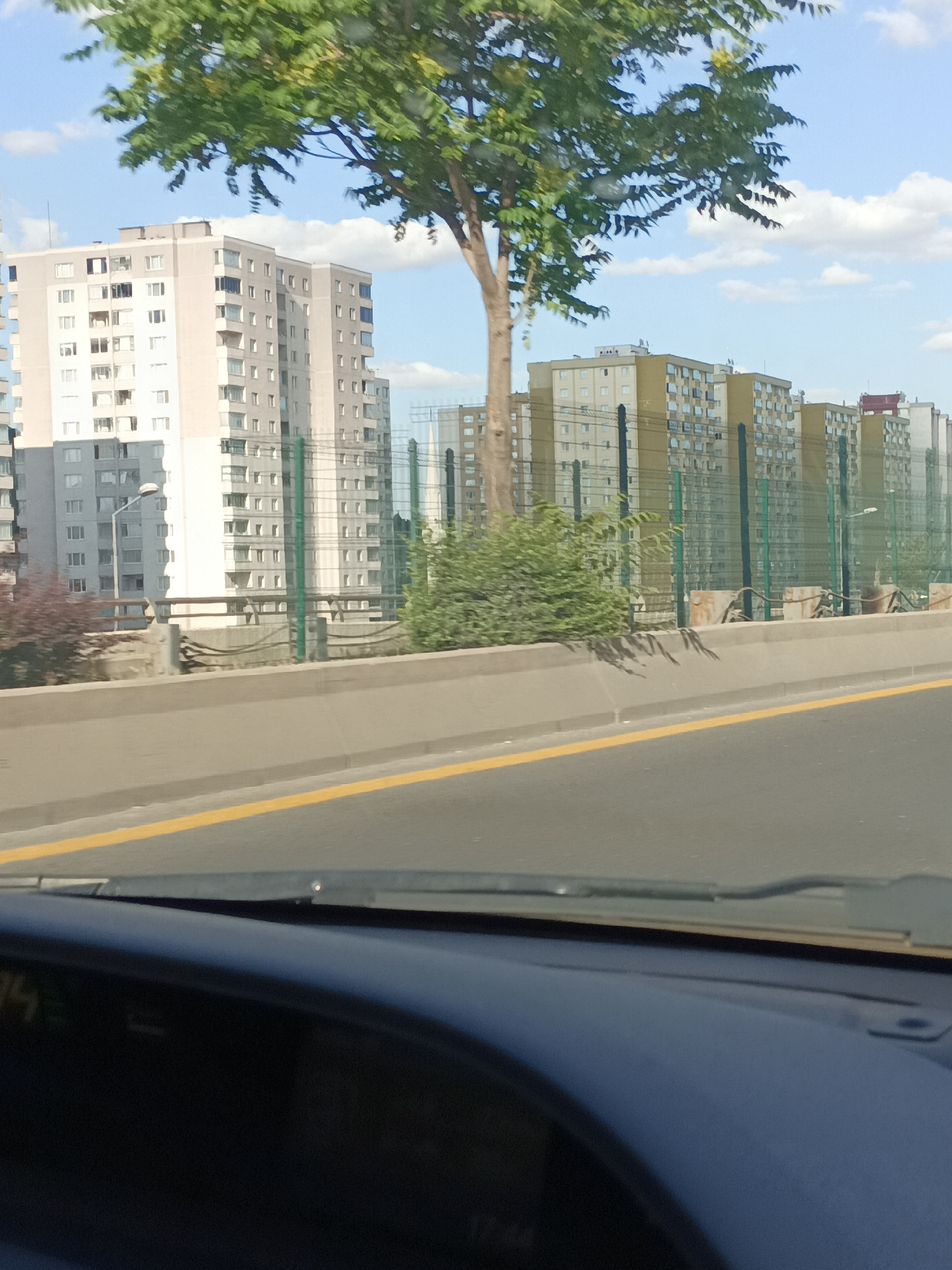 This media file depicts a roadside view of multiple apartment buildings and a tree with green leaves against a blue sky with scattered white clouds. The perspective appears to be from inside a vehicle, as the edge of a dashboard and windshield are visible in the foreground. A concrete barrier with a yellow line separates the road from the area with the buildings.
The apartment buildings are modern, mid-rise structures, some appearing to be light-colored and others with yellow facades. They are arranged in a row, with some distance between them. A green metal fence is visible in front of some of the buildings. A street lamp is also present.
The time of day appears to be daytime, given the bright lighting and shadows. The weather seems pleasant, with a clear blue sky. The context provided places the scene in Mamak, Turkey. There is no visible text in the image that provides any specific information. The overall impression is that of a residential area in an urban or suburban setting.