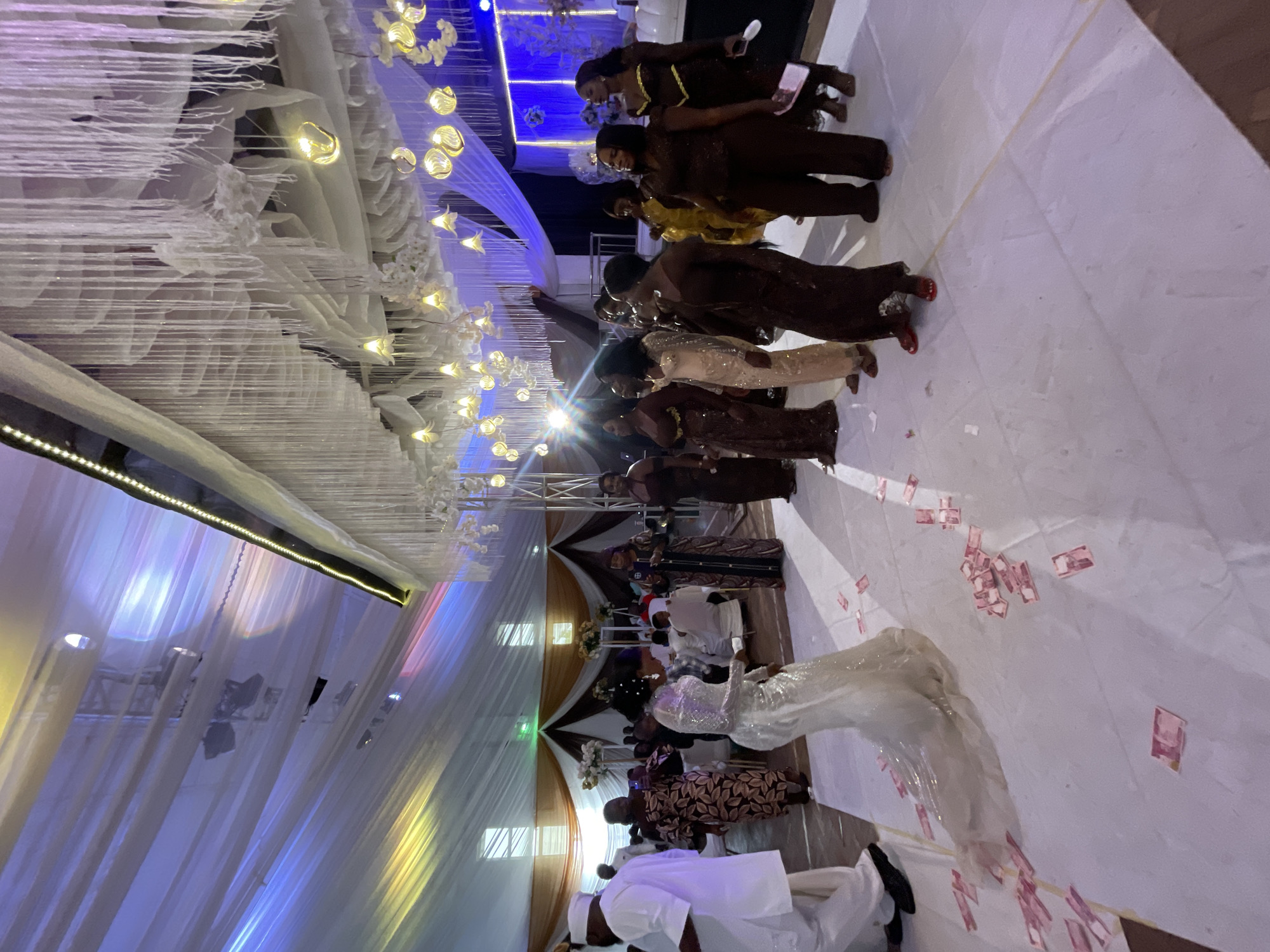A wedding ceremony in Unknown, Unknown, shows a bride and groom with guests in an indoor setting. The interior is decorated with white fabric draping from the ceiling, enhanced by light fixtures and string lights. Along the aisle are several people, presumed to be wedding guests, standing to the side. Currency notes are scattered along the aisle on the floor.