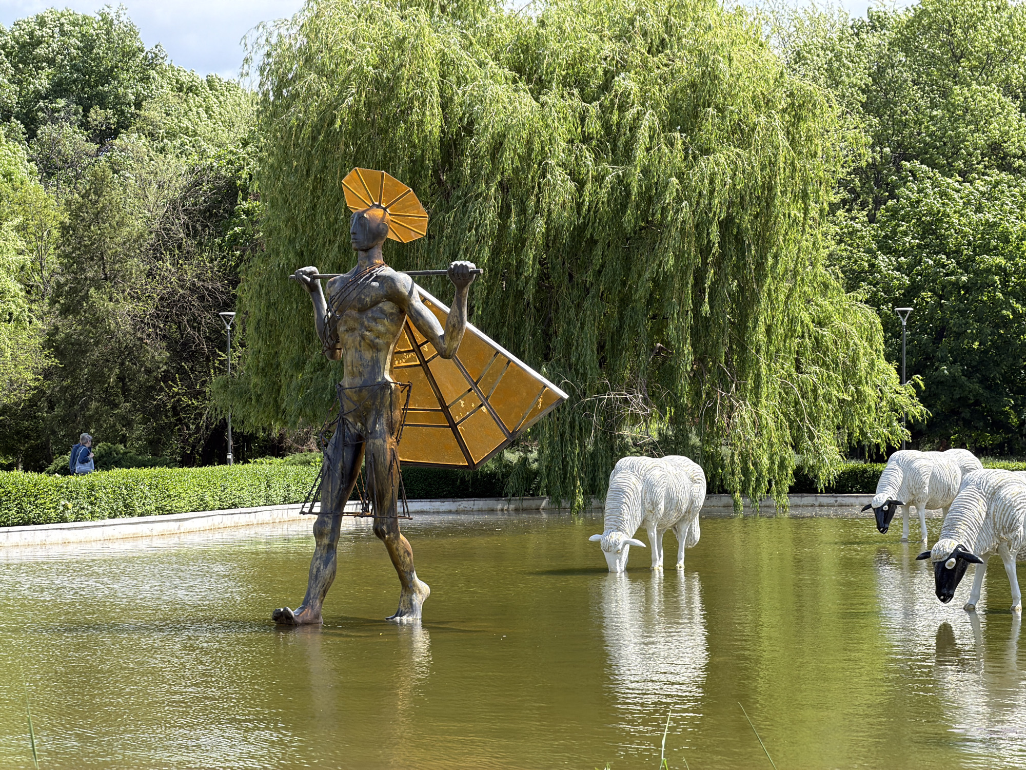 The image depicts a statue of a human-like figure carrying a yoke, in the waters of an artificial pond in Borisova Gradina park in Sofia, Bulgaria. The sculpture is accompanied by the statues of three sheep, standing in the pond. The water's surface reflects the statues and surrounding greenery. Lush green trees surround the pond, with a grassy bank and hedge visible in the background. A person is visible beyond the hedge.
