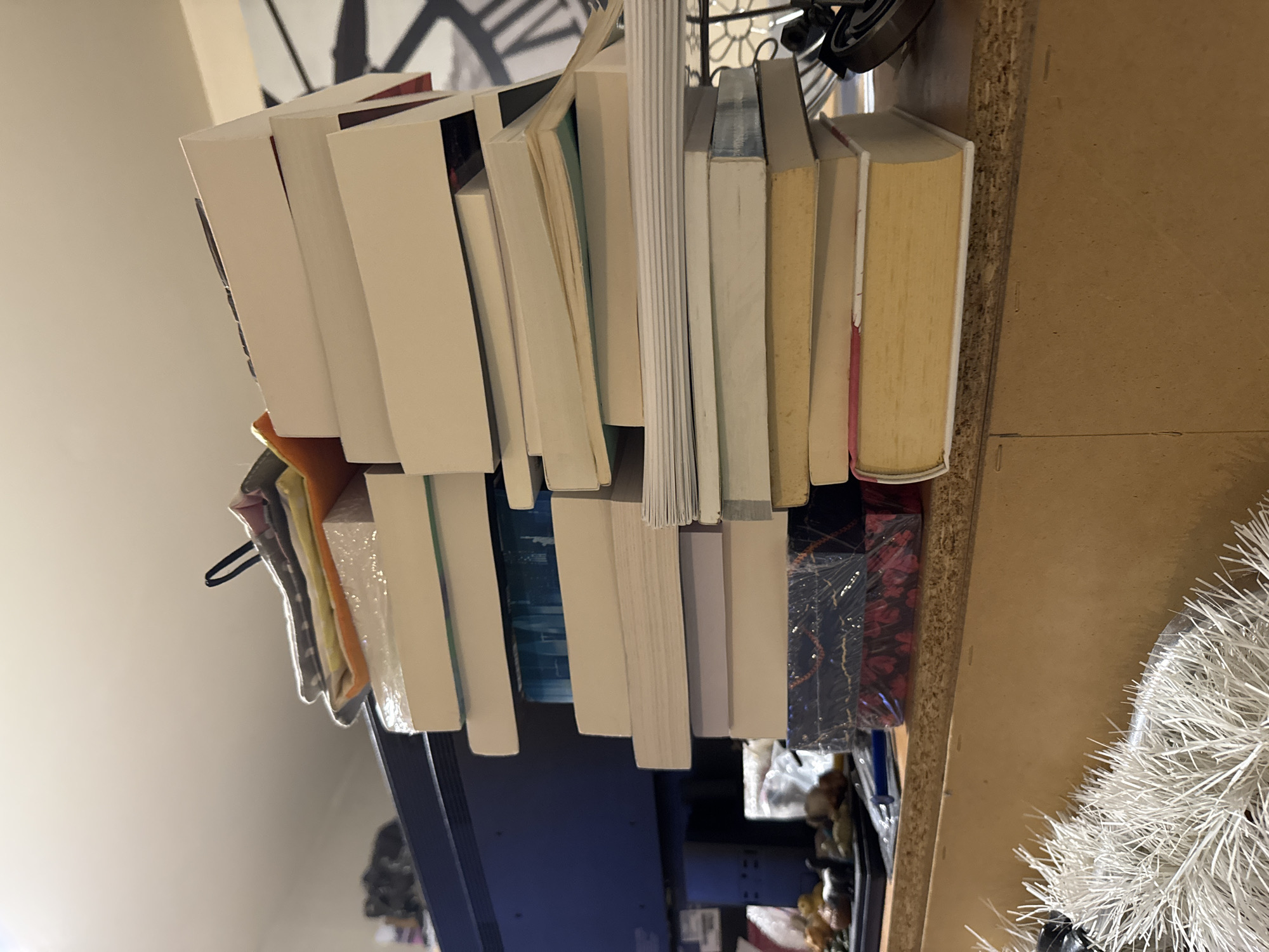 The image depicts a stack of books placed haphazardly on a surface, likely a shelf or table. The books vary in size and color, with a mix of white, cream, and light blue covers. There are also some decorative items visible, including what appears to be a white decorative plant and some wrapped items. The background suggests an interior setting with a clock and a plain wall. The location of the image is not evident, and there is no information to suggest it is taken in Sofia, Bulgaria.