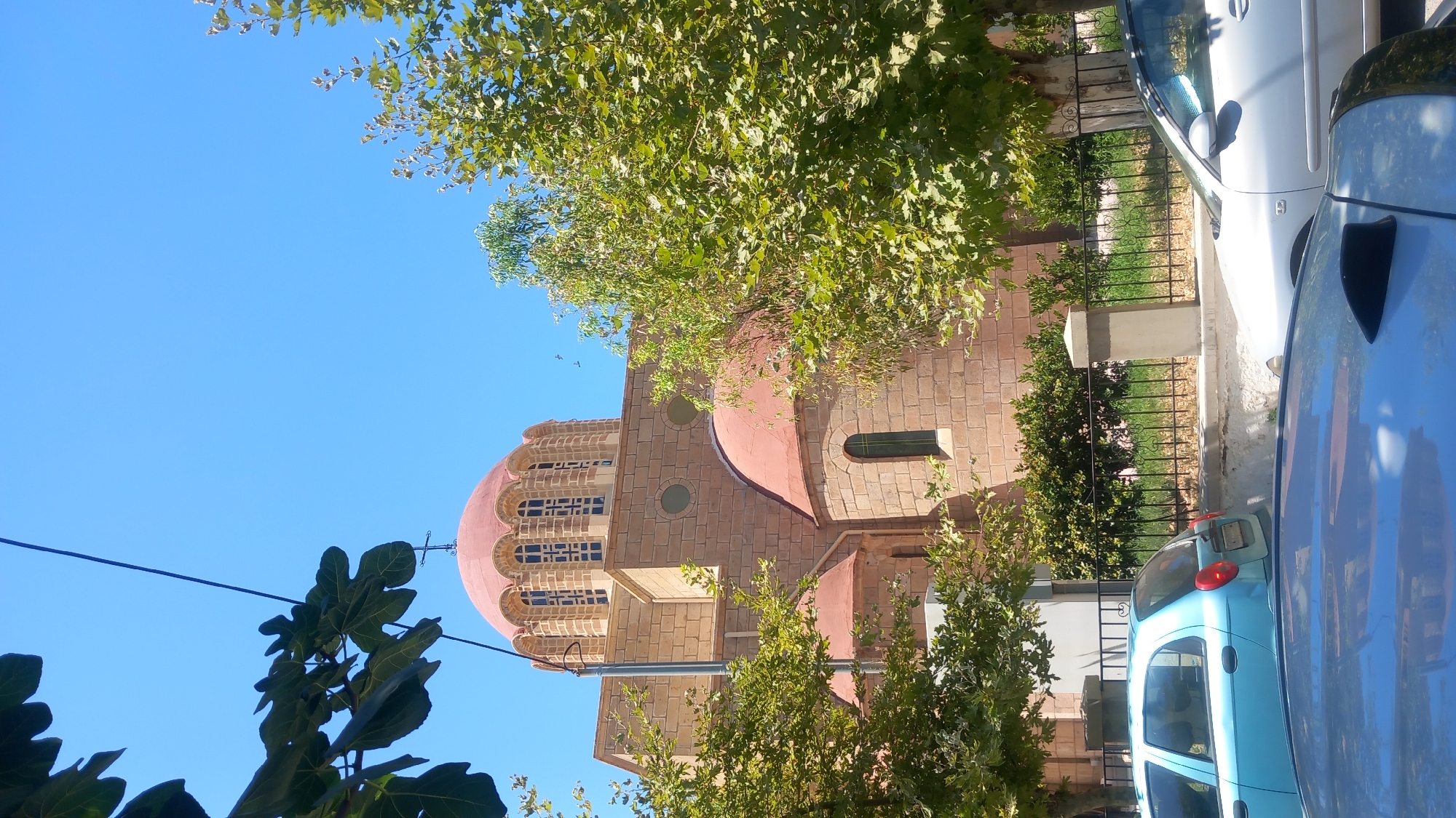 The image captures a sunny day in the city of Kos, Greece, showcasing a portion of a church with distinctive red domes and stone brick construction. A prominent large dome with arched windows topped by a cross sits above a smaller dome, both exhibiting a reddish-orange hue. The church's facade is constructed from light-colored stone blocks. Lush green trees with dense foliage frame the building, partially obscuring it from view. In the foreground, the tops of several cars are visible. A light blue hatchback is parked on the left, and a silver sedan is parked to its right. A black fence with decorative scrollwork runs along the front of the church property, separating it from the street and parked vehicles. The sky above is a clear, vibrant blue.