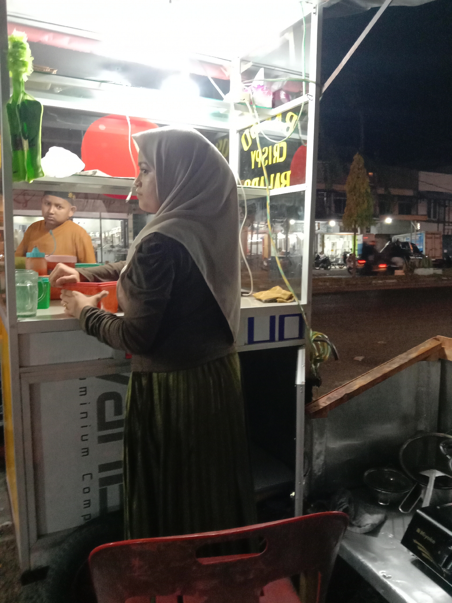 The image shows a woman in a hijab standing in front of a food cart on a street in Lhokseumawe, Indonesia. The food cart is brightly lit and displays items like a red balloon, various food containers, and a sign with the words "CRISPY" and "BALADO" written on it. A boy wearing an orange shirt is visible inside the cart, behind the woman. The street outside the food cart is dark, with buildings and streetlights visible in the background. There are also vehicles parked along the street. A red chair is in the foreground.