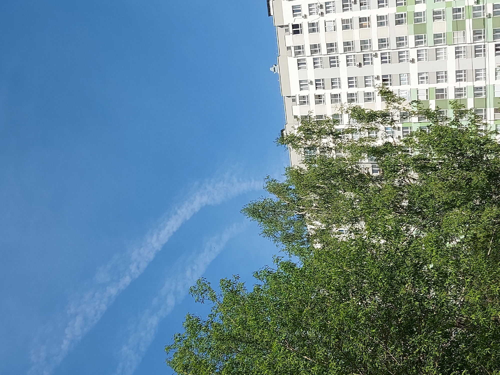 A low-angle shot captures the exterior of a multi-story residential building in Izhevsk, Russia. The building, primarily white with interspersed sections of light green, features rows of windows and multiple air conditioning units mounted on the facade. Green trees fill the right lower portion of the frame, partially obscuring the lower levels of the building. The sky is a clear blue, with contrails from a plane visible on the left side.