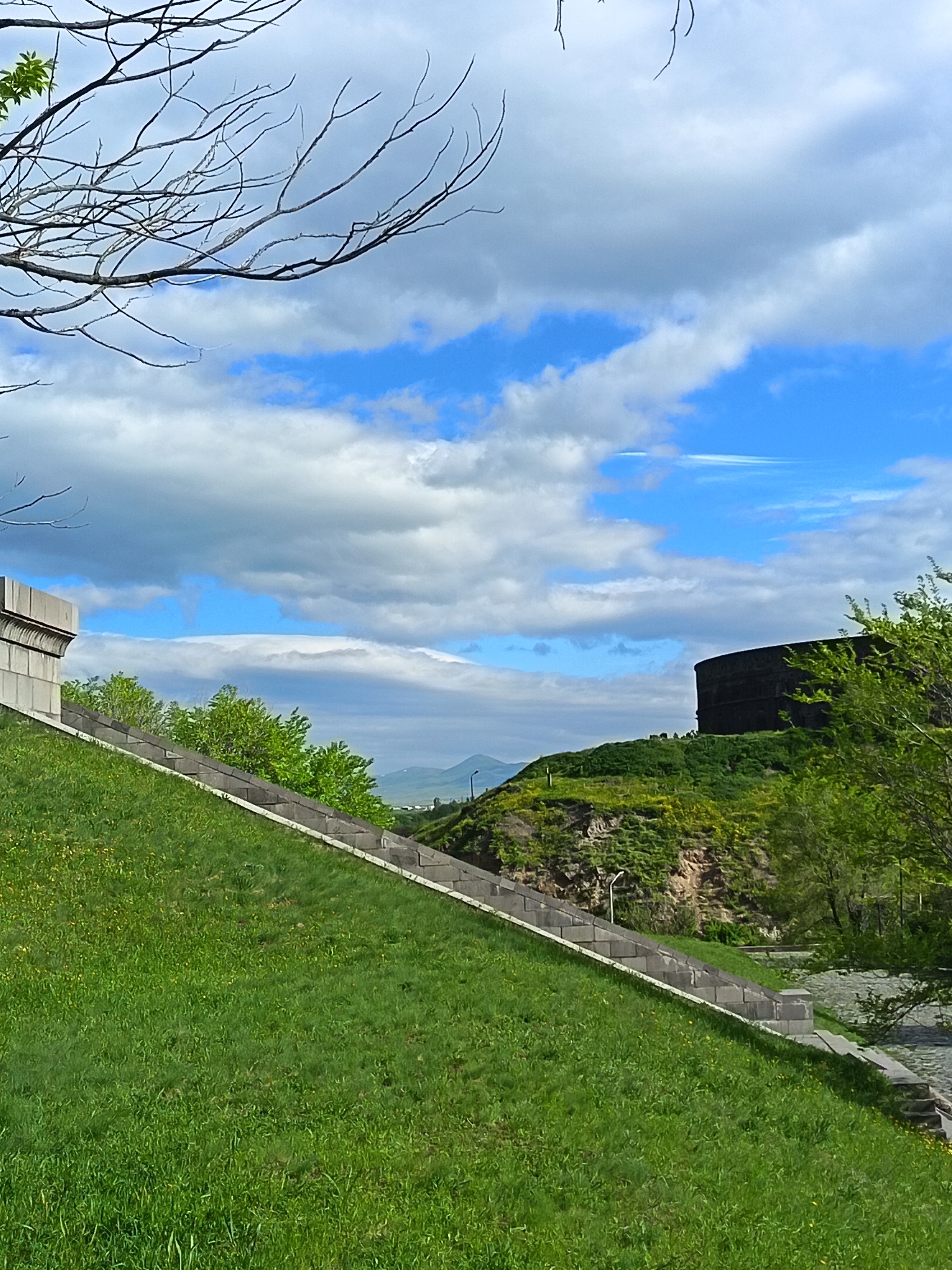 The image shows a cityscape featuring green spaces and architectural elements under a partly cloudy sky. In the foreground, a grassy slope is bordered by a gray stone stairway. Trees and greenery are scattered along the slope and to the right of the frame. In the middle ground, a hill is topped by a dark, cylindrical structure, possibly a building or monument. Further in the distance, a mountain can be seen on the horizon through a break in the buildings. The sky is blue with white clouds, and bare tree branches are visible in the upper left corner.