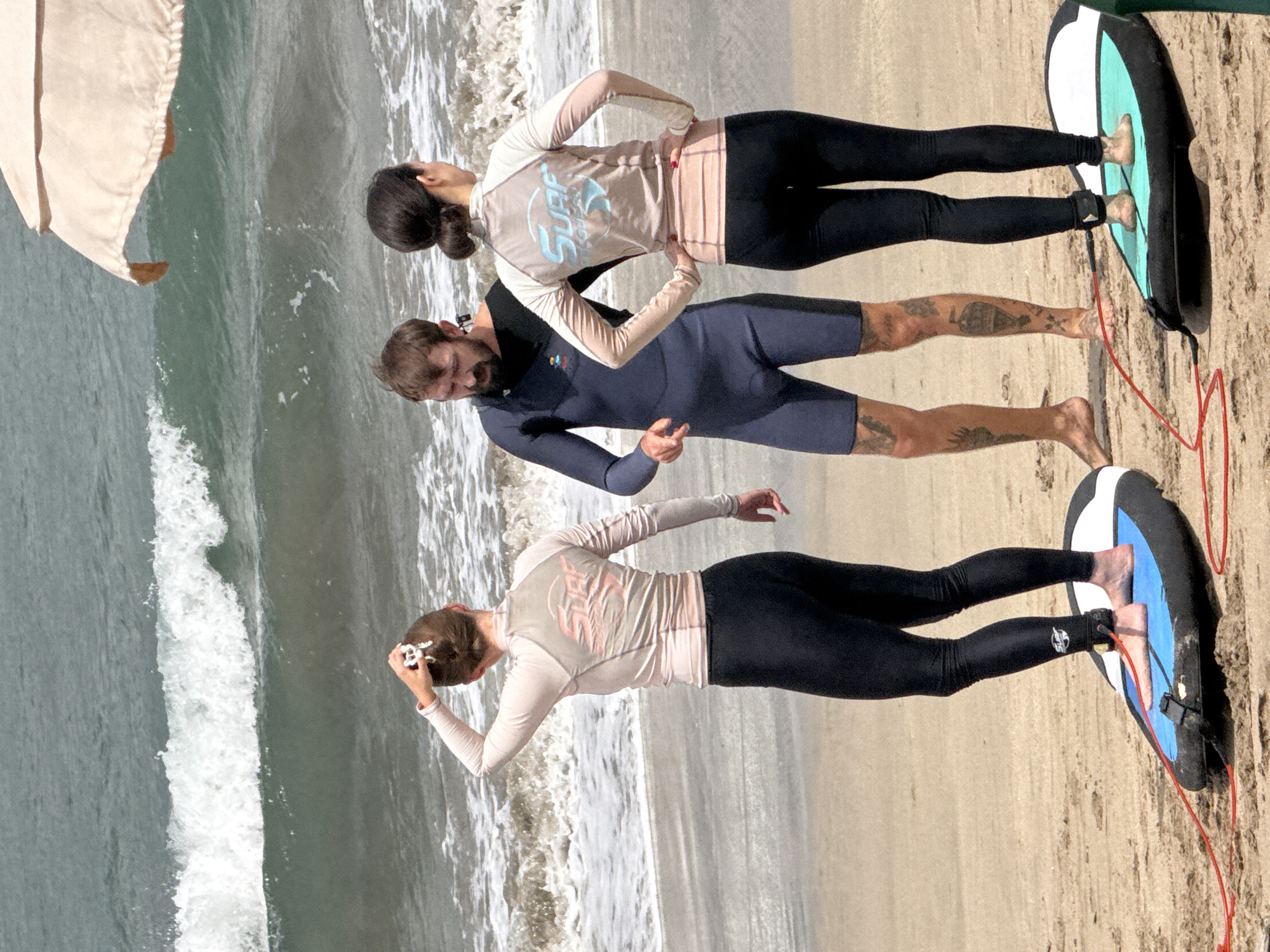Three individuals stand on a sandy beach in Kuta, Indonesia, with the ocean and waves in the background. A light-colored umbrella stands to the right. Two women in surf attire, one with her hair up and the other with her hair in a bun, face the ocean. A man in a wetsuit stands between them, also facing the ocean. The two women are each standing on a surfboard. The man is barefoot, and the women have their feet on their boards. The scene appears to be a surfing lesson or a group preparing to surf.