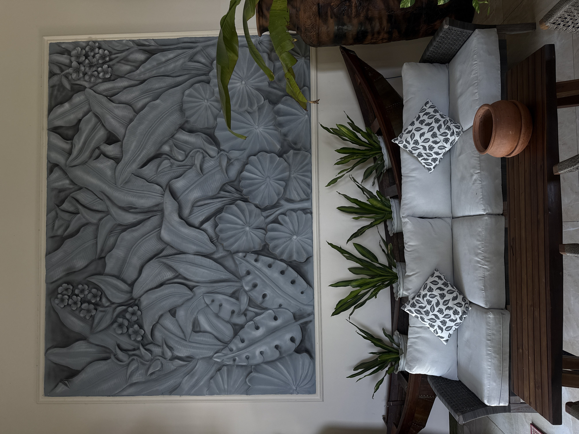 The image shows an indoor living area, likely a hotel lobby or restaurant in Bac Nha Trang Ward, Vietnam. A light-colored, three-cushion sofa is placed in front of a large, grey bas-relief artwork depicting tropical foliage and flowers. Two decorative pillows with a black leaf pattern sit on the sofa. In front of the sofa is a dark wooden coffee table with an unglazed terracotta pot and saucer on it. Behind the sofa, a row of five potted green plants are visible, and behind them, a dark wooden canoe-like object is propped against the wall. To the right of the sofa and plants, a large, dark, textured ceramic vase stands, with a large green plant with broad leaves extending from it. The wall behind the artwork is painted a light, neutral color. The flooring appears to be tiled. No people are present in the scene.