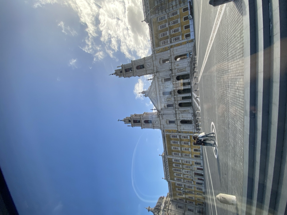 The image depicts the Mafra National Palace in Mafra, Portugal, under a clear blue sky with scattered white clouds, predominantly on the right. The sun is bright, casting shadows across the foreground.
The central subject is the grand Baroque complex of the Mafra National Palace, featuring a prominent white stone basilica with two symmetrical bell towers. Each tower is topped with a weather vane and displays a clock face. The basilica's facade includes multiple columns, arches, and sculptural elements, with a wide set of steps leading up to its main entrance. Flanking the central basilica are two extensive wings of multi-story buildings, characterized by their yellow-ochre facades and numerous windows with gray detailing. Scaffolding is visible on the far right section of the palace's right wing, indicating ongoing work.
In the large, paved square (Terreiro D. João V) in front of the palace, three individuals are standing together near the center. They are positioned within a circular marking on the dark grey cobblestone-like pavement, which features lighter contrasting lines forming a geometric pattern. The individuals are casually dressed. A small white utility vehicle is parked near the base of the palace's right wing.
The foreground shows several dark, horizontal steps, suggesting the image was captured from a slightly elevated position, possibly through glass, as faint reflections are visible across the lower portion of the frame. A subtle, curved white contrail is visible in the upper left section of the blue sky.