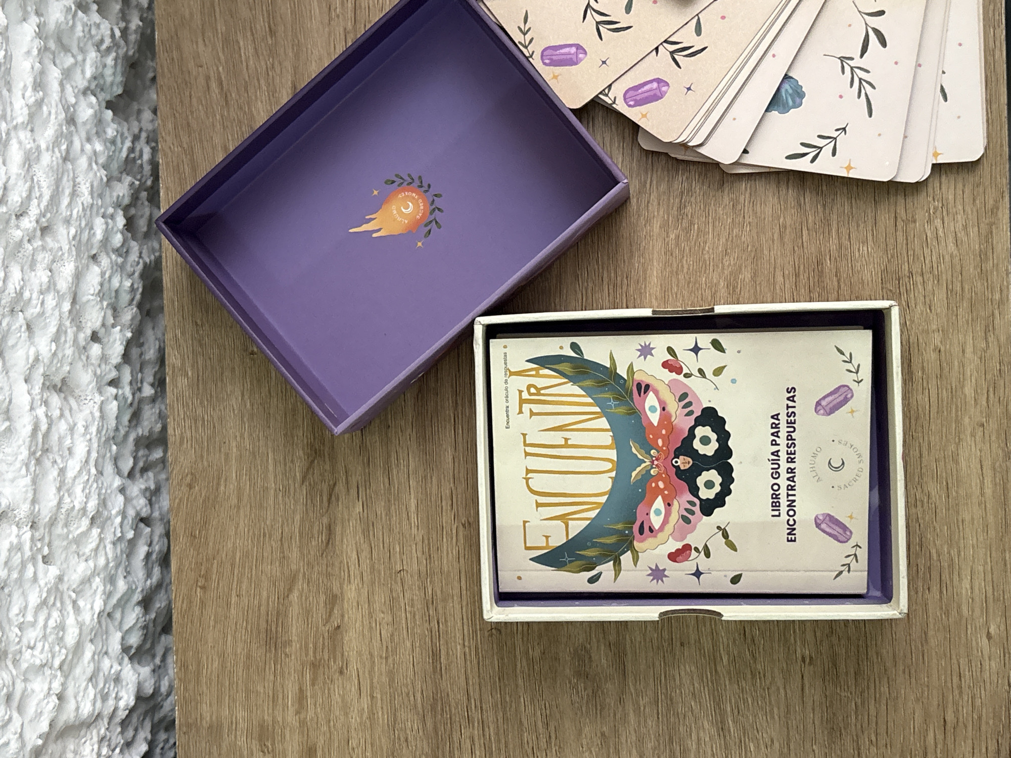 An overhead shot displays a spiritual or self-help product set arranged on a light brown wooden surface, with a textured white wall visible on the left side of the frame. The scene is set indoors, likely in Puebla, Mexico.
The central items include an open, rectangular purple box. Its inner lid features a vibrant emblem of a stylized comet, with a circular design that reads "ALHUMO SACRED SMOKES" and "ALHUMO". Adjacent to this, to the right, is a smaller white box containing a guide book.
The book's cover is prominently displayed, featuring the large title "ENCUENTRA" (Find) and the text "LIBRO GUÍA PARA ENCONTRAR RESPUESTAS" (Guide Book to Find Answers), with a smaller line stating "Encuentra: oráculo de respuestas" (Find: oracle of answers). The cover art is elaborate, showcasing a crescent moon, a detailed butterfly with colorful wings, scattered stars, purple crystals, and botanical motifs. A small circular logo on the book also repeats "ALHUMO SACRED SMOKES."
To the upper right, partially fanned out, is a stack of oracle or tarot cards. The visible card backs feature a light background decorated with purple crystal illustrations, small stars, and delicate green leafy branches, complementing the mystical and natural theme of the set. The overall aesthetic is cohesive, suggesting a tool for introspection or seeking guidance.