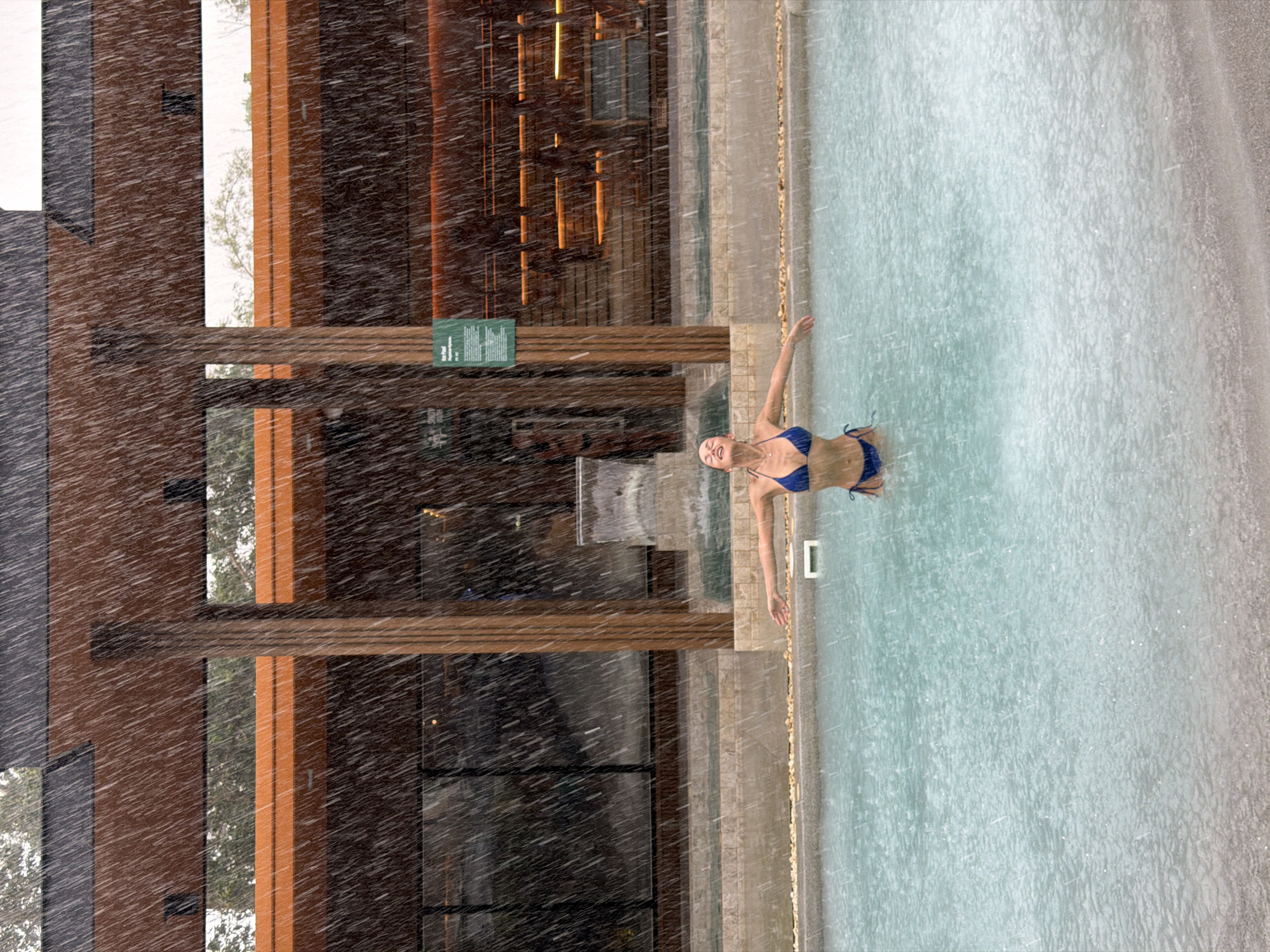 A woman in a blue bikini lies supine in a pool during a heavy rainstorm. The pool is bordered by light-colored stonework. Behind the pool is a building with a wood frame and glass windows. The location is in the city of Si Sunthon, Thailand.