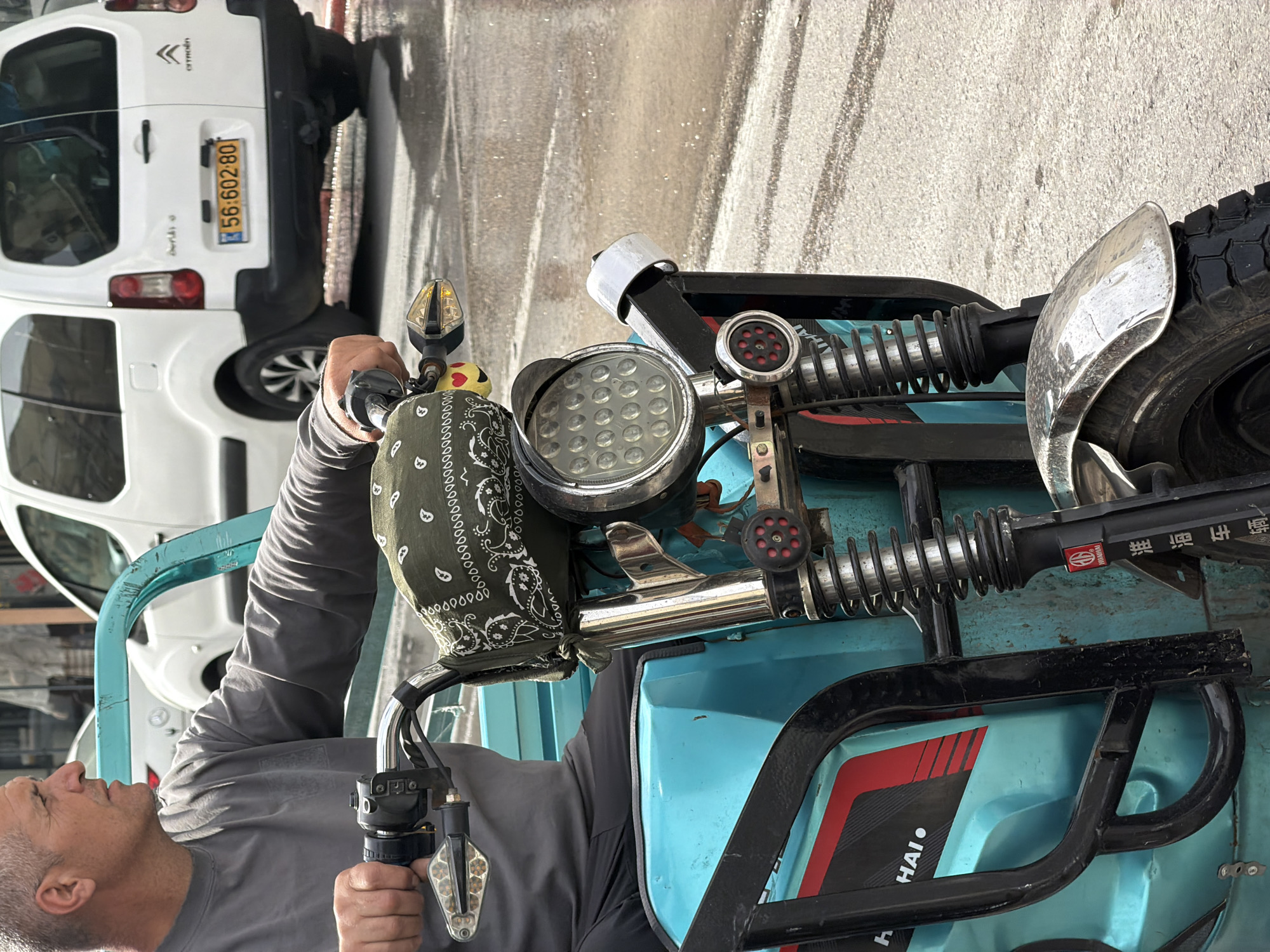 The image is an unusual, high-angle shot of a man sitting on a blue tricycle with a white car parked nearby. The man has short, gray hair and is wearing a gray shirt. His hands are gripping the handlebars of the tricycle, which has a black and white bandana tied to it.
The tricycle is light blue with black trim and features a large headlight. The white car in the background is a Citroen, with the license plate number 56:602-80 visible.
The letters 'HAI' are visible on the blue body of the tricycle. The location is most likely in the city of Tel Aviv, Israel, given the Israeli license plate on the Citroen. The setting appears to be an urban street.