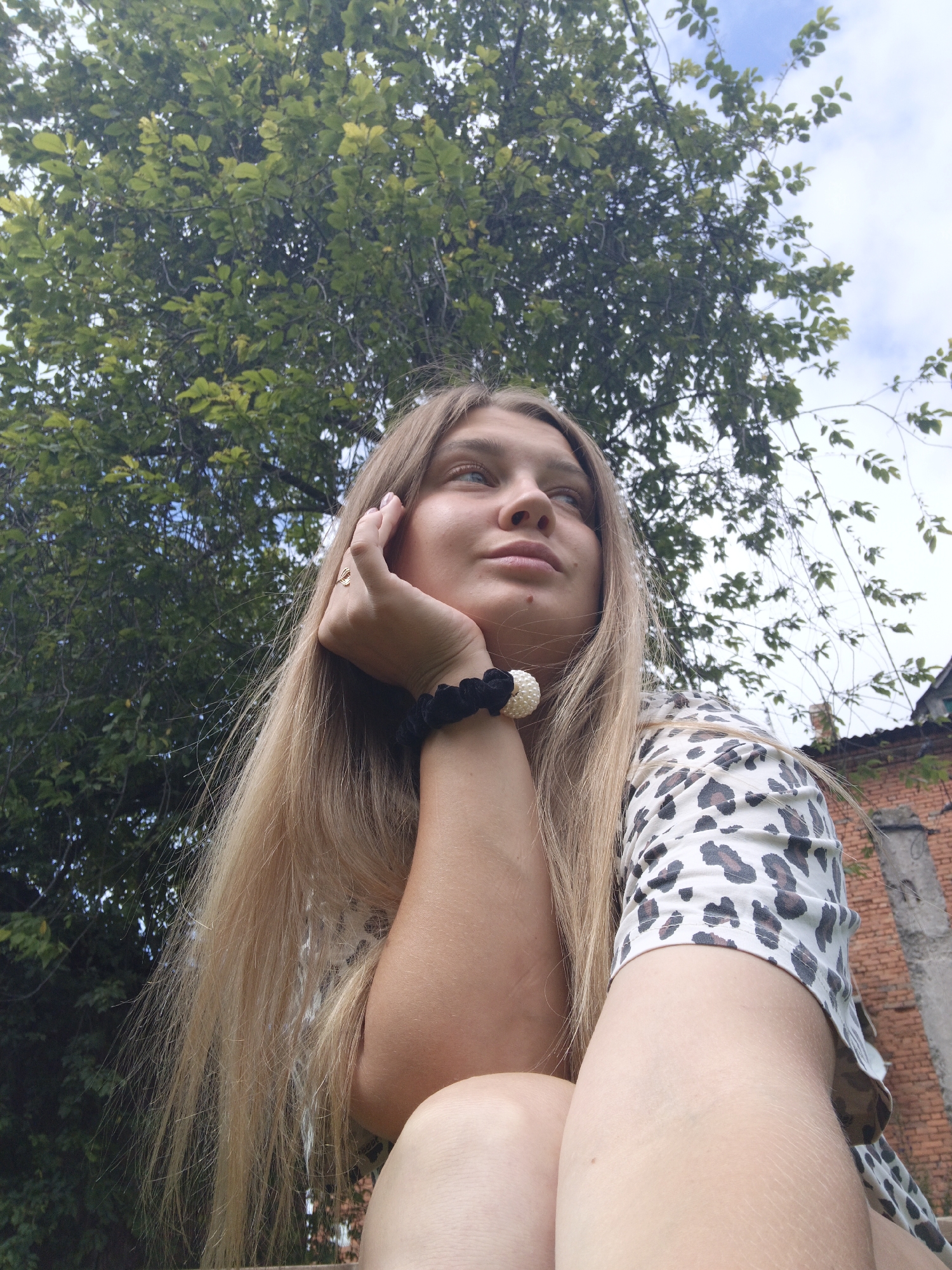 A young woman with long blonde hair and a leopard print shirt is posing outdoors. She is resting her chin on her hand, and her gaze is directed upwards and to the side. She is wearing a black scrunchie with white beads on her wrist. The background consists of green trees and a cloudy sky. Partially visible to the right is a brick building. The scene appears to be in Medvedyovka, Russia.