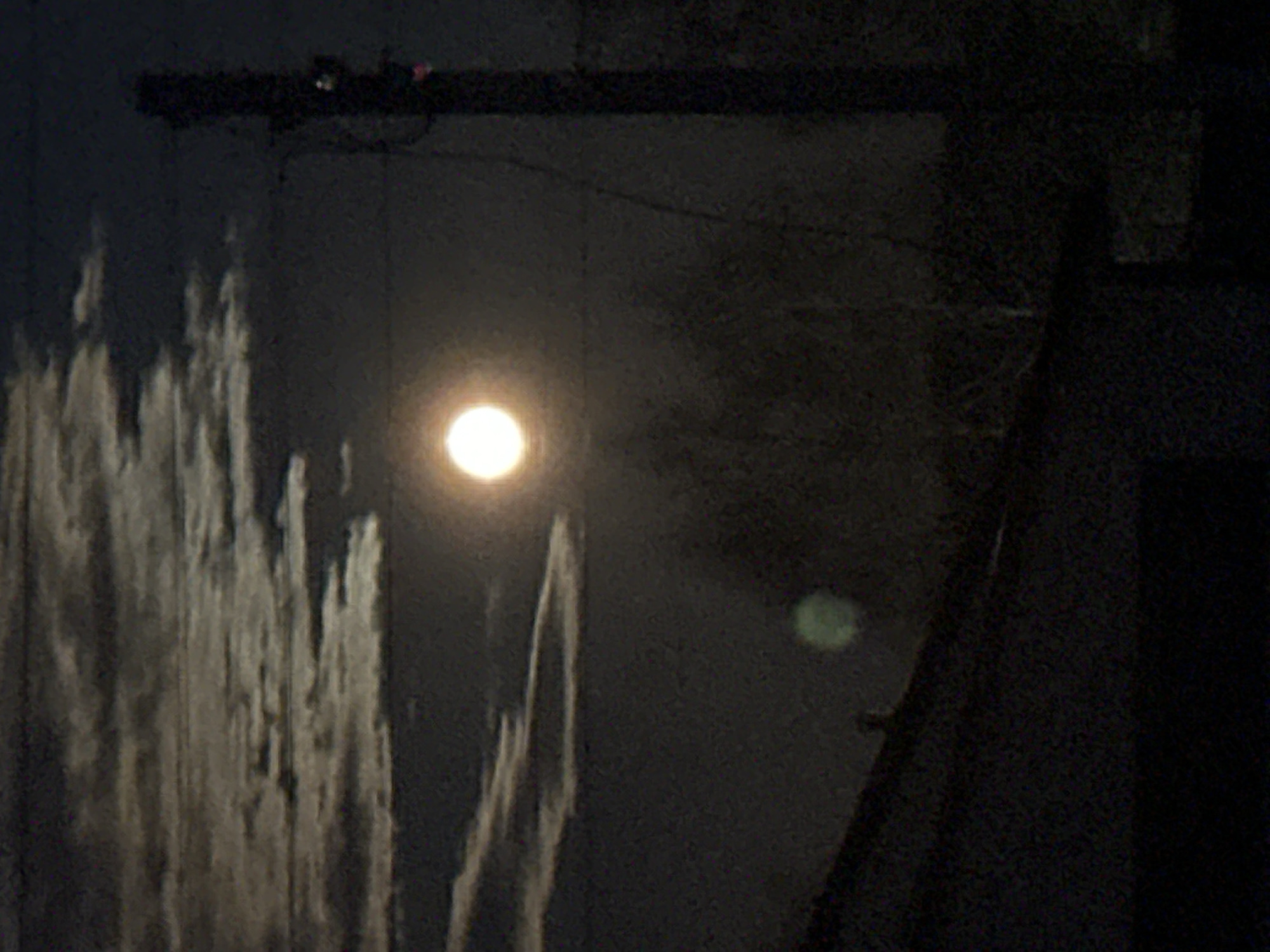 The image depicts a dark, possibly nighttime scene in Kharnavy, Russia. In the center is a bright light source, likely a street lamp or the moon, surrounded by dark sky. To the left are vertical streaks or reflections of light, possibly from wet surfaces or a body of water. A dark, horizontal structure is visible at the top, possibly a utility pole or beam with attached devices. A dark, angled shape is on the right, obscuring some of the background, possibly a building corner or roofline. A faint, greenish light is also present to the right of the central light source.