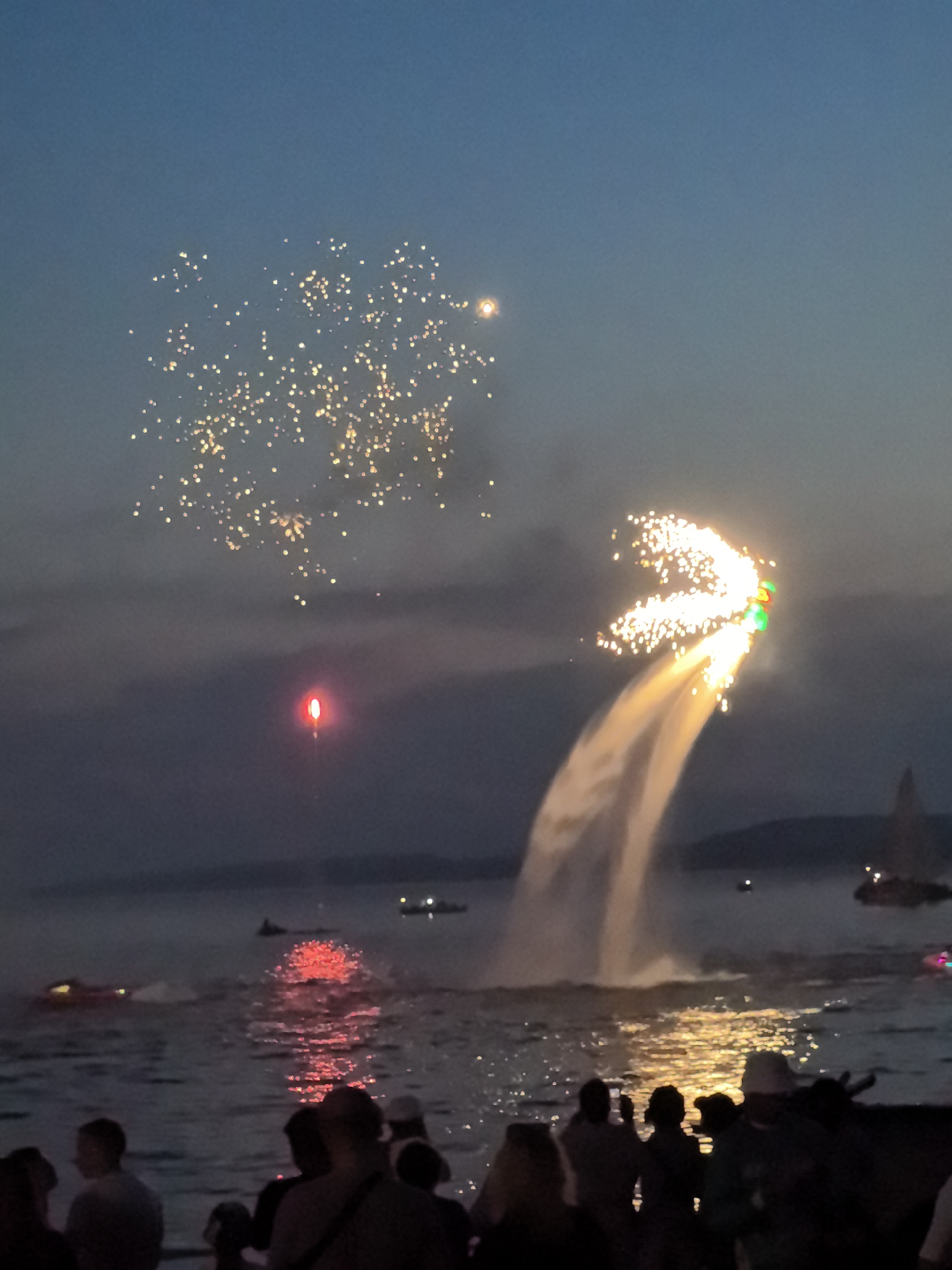 This image depicts a waterfront scene at dusk, likely in Osa, Russia, during a pyrotechnic display. In the sky, a cluster of golden fireworks is visible, along with a single red flare or light source. To the right, a person on a hydro-powered jetpack is launching water and sparks into the air, creating a dramatic effect. Several boats are present on the water, including a jet ski on the left and what appears to be a sailboat in the distance. The foreground shows a silhouetted crowd of people gathered along the waterfront, observing the spectacle. The sky is a gradient of deep blue and dusky grey, with the water reflecting the lights from the fireworks and the jetpack.