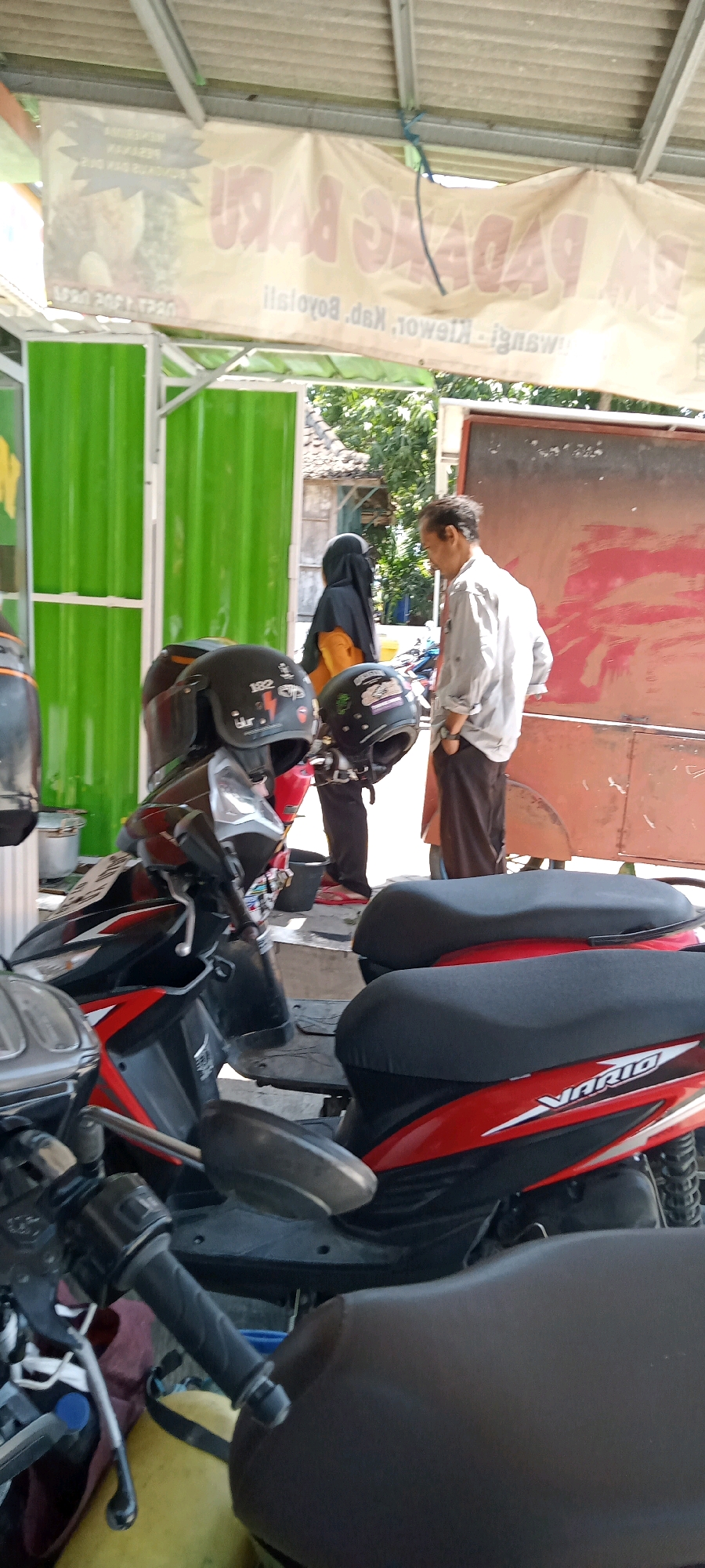 The image depicts an outdoor scene likely in Desa Karangmojo, Indonesia. In the foreground, two black motorcycle seats are visible, one with a red and black patterned side panel labeled "VARIO". Several motorcycles are parked, with two black helmets prominently displayed on the handlebars of the nearest one. A man in a light-colored shirt and dark pants is standing and looking down, while a person wearing a black hijab over an orange garment stands to his left. Behind them, a large, faded banner with red text is stretched across the top of the frame, appearing to advertise a business. To the left, a bright green corrugated metal wall forms a barrier, and beyond that, trees and a portion of a building are visible. The overall environment suggests a roadside establishment or parking area.