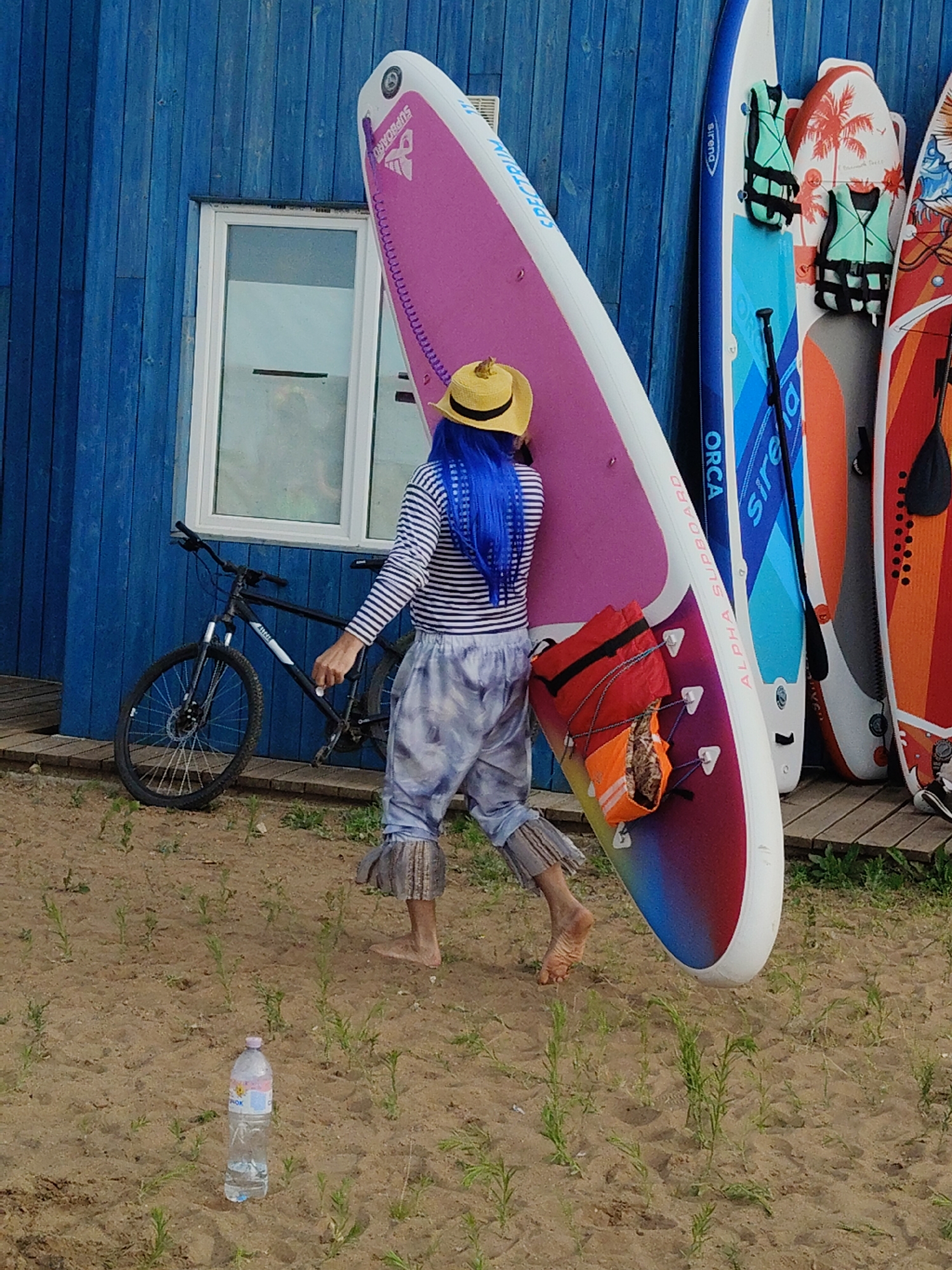A person with bright blue hair and a straw hat is carrying a pink and rainbow-colored stand-up paddleboard on a sandy beach. They are wearing a striped long-sleeved shirt and patterned pants. To the left of the person is a black bicycle. In the background, there is a blue wooden building with a window and several other stand-up paddleboards leaning against it. A bottle of water is visible on the sand in the foreground.