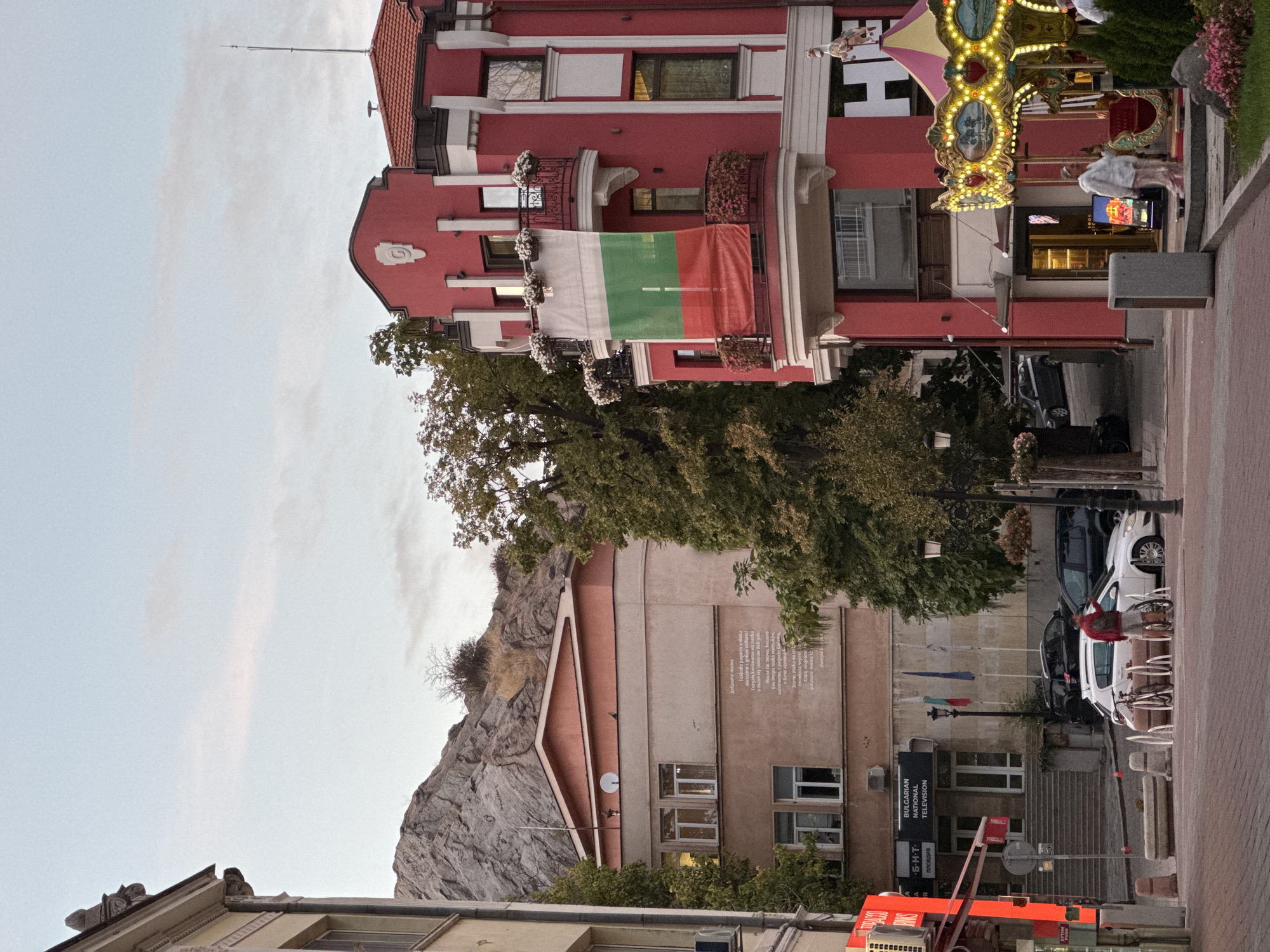 In Plovdiv, Bulgaria, a scene unfolds with a striking pink building featuring a balcony adorned with flowers and a Bulgarian flag. Across the street, a beige building displays text, possibly poetry, and is backed by a rocky hill. A carousel with illuminated panels and a decorative horse stands to the right. A man with a backpack stands beside a bicycle in front of a white car, while other vehicles are parked nearby. The environment suggests a public space, possibly a plaza or street, with trees lining the sides. The sky is overcast, indicating twilight or a cloudy day.
