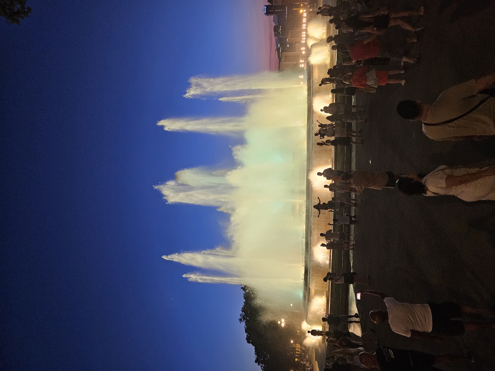 The image captures the Magic Fountain of Montjuïc in Barcelona, Spain, at twilight. The sky is a deep blue, transitioning to a soft pink and orange hue near the horizon. The fountain is illuminated with bright, yellowish-green light, creating a luminous spectacle with multiple jets of water reaching high into the air.
In the foreground and midground, numerous people are gathered, silhouetted against the fountain's glow. Many are standing, facing the fountain, with some holding up their phones to take pictures or videos. A few individuals are seen walking. The ground appears to be a dark, paved area.
Behind the fountain, trees line the background, and further in the distance, buildings are visible, with one bearing an "Allianz" logo. The overall scene depicts a public gathering to witness a nighttime water and light show.