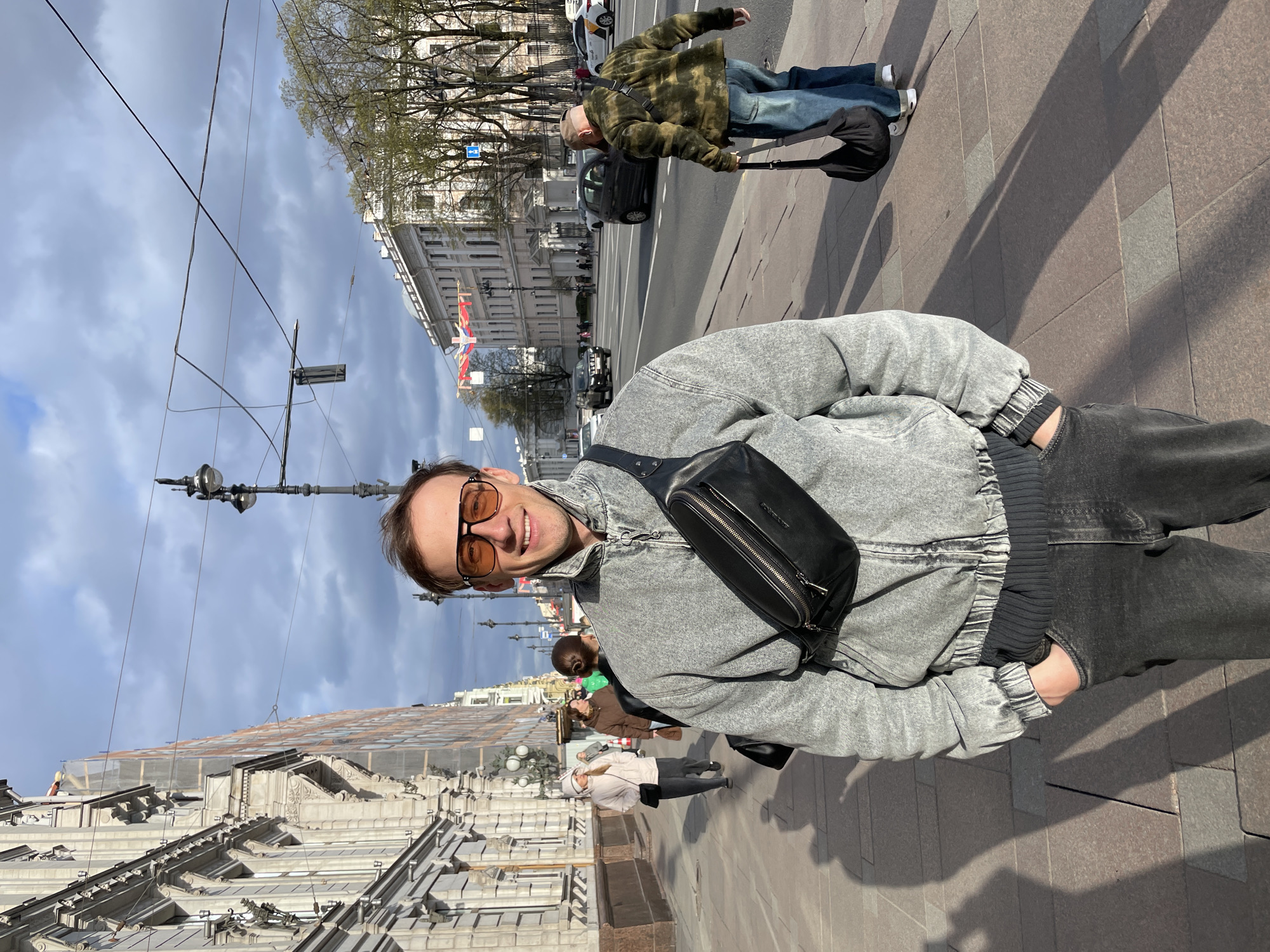 A young man poses for a photo on a sunny city street, likely in Saint Petersburg, Russia, based on the context. He is the central subject, smiling and looking directly at the camera, wearing amber-tinted sunglasses, a light grey, acid-washed denim-style bomber jacket, and dark jeans. A black fanny pack is slung across his chest. His hands are casually in his pockets.
The setting is an urban environment with classic European architecture. In the background, a wide street is visible with cars and several pedestrians. On the right side of the street, there are large, ornate buildings, some adorned with flags displaying red, white, and blue stripes (suggesting Russian flags). One building on the left side of the street appears to have construction netting on its upper levels. Overhead wires, possibly for trams or trolleybuses, crisscross the sky.
The weather appears to be pleasant with clear blue sky interspersed with light clouds. Bright sunlight casts distinct shadows on the paved sidewalk, indicating mid-morning or afternoon. Trees with budding green leaves line the street, suggesting spring or early summer. Another man with a camouflage jacket and jeans is visible further down the street, pulling a luggage bag. Other pedestrians are scattered along the sidewalk. The overall scene captures a lively moment in a bustling city. There is no visible legible text in the image.