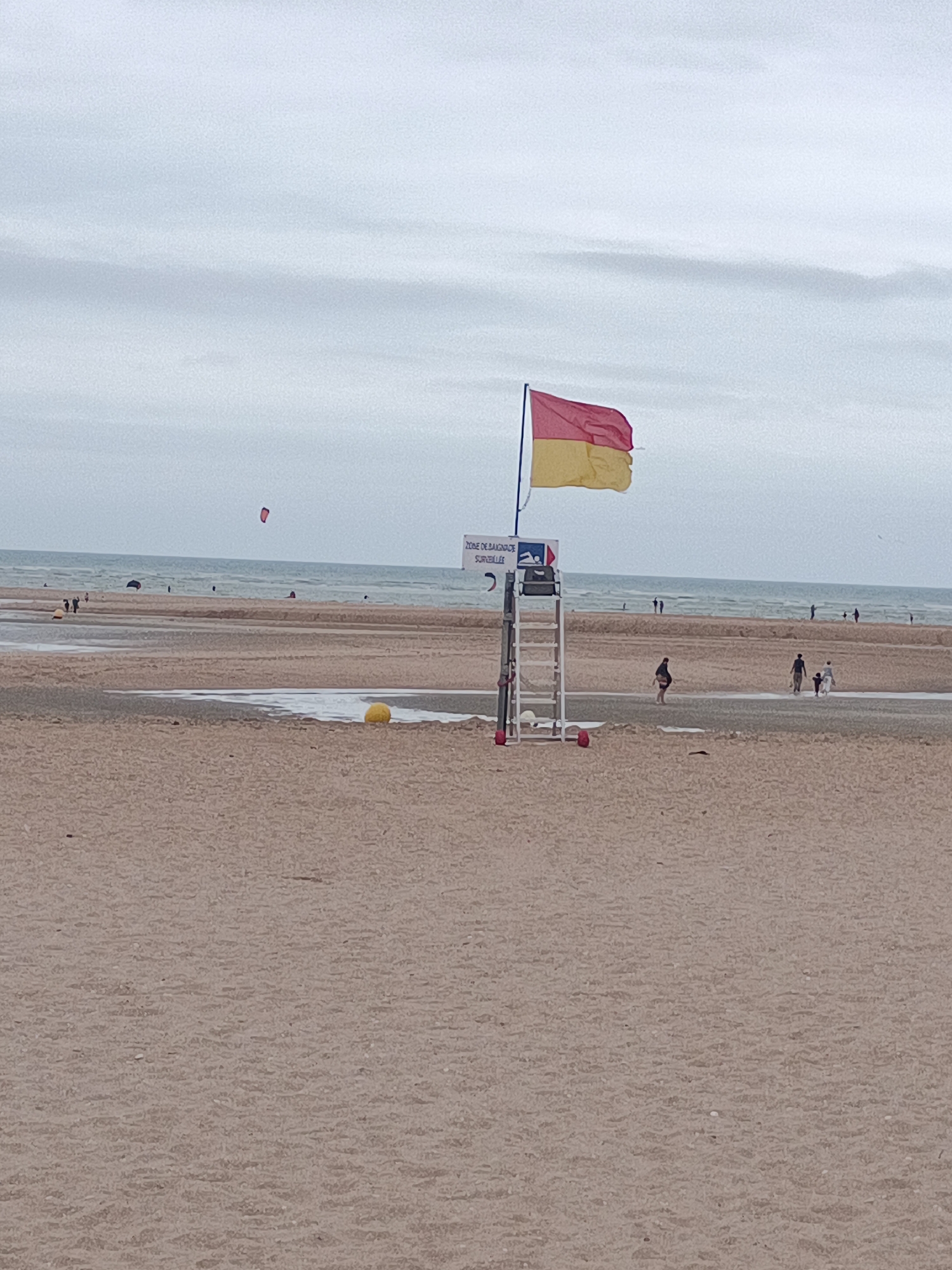 The image shows a beach scene in Houlgate, France, under a cloudy sky. In the foreground, there is a sandy beach with some shallow water. In the mid-ground, a lifeguard tower stands with a red and yellow flag flying from it. There are a few people walking on the beach and in the water. In the background, the sea is visible, with some distant figures and a kite surfer. A yellow buoy is visible in the shallow water to the left of the lifeguard tower.