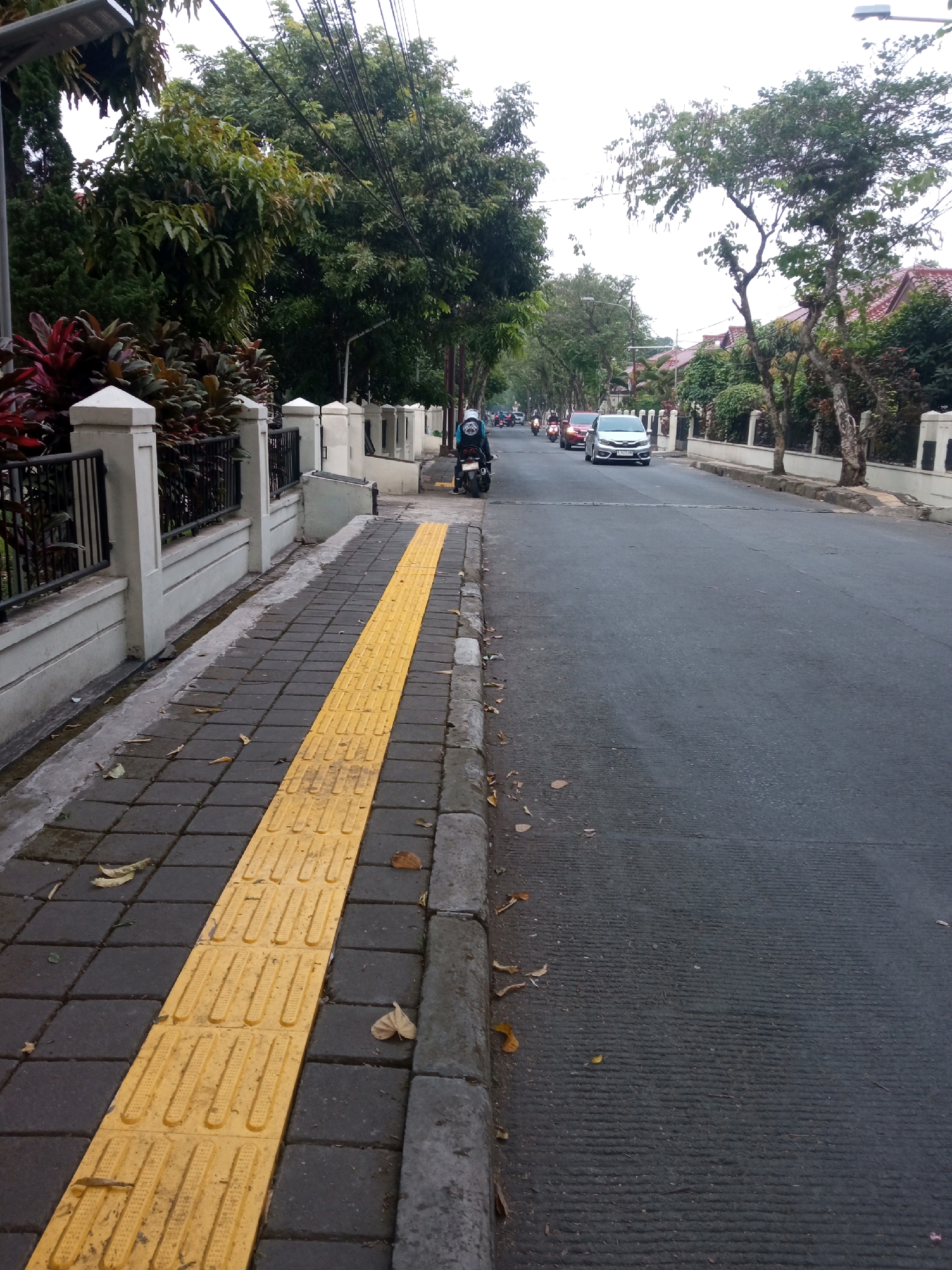 The image shows a street scene in Bandung City, Indonesia. On the left side of the image, there is a sidewalk paved with dark gray square tiles, with a raised yellow tactile paving strip running down its center. A black metal fence with white pillars lines the edge of the sidewalk, separating it from a residential area with lush green trees and houses. On the right side of the image, a gray asphalt road is visible, with a few cars and motorcycles driving on it. The sky is overcast, suggesting a cloudy day. The overall scene depicts a typical urban street in Indonesia, with a focus on pedestrian infrastructure and the presence of vehicular traffic.