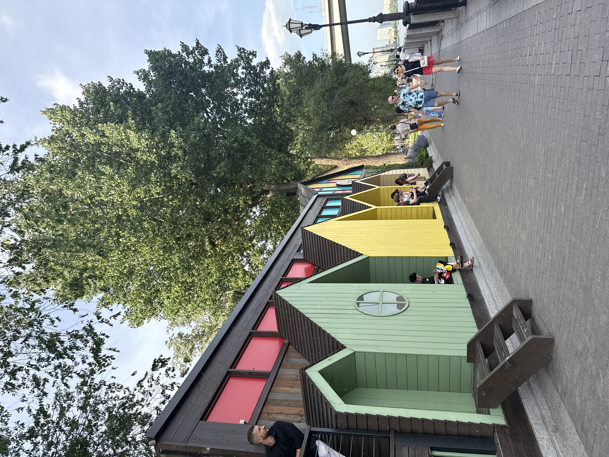 The image depicts a series of colorful, stylized A-frame structures located in Rostov-on-Don, Russia. The structures are painted in different colors including black and red, green, yellow, and light blue. Several people are visible in and around the structures, some sitting inside the cubicles and others standing nearby. A paved walkway stretches along the side of the structures, and several people are walking along the walkway. A large tree with dense foliage stands adjacent to the structures. A bridge is seen in the background. The sky is clear, and the overall lighting suggests it is daytime.