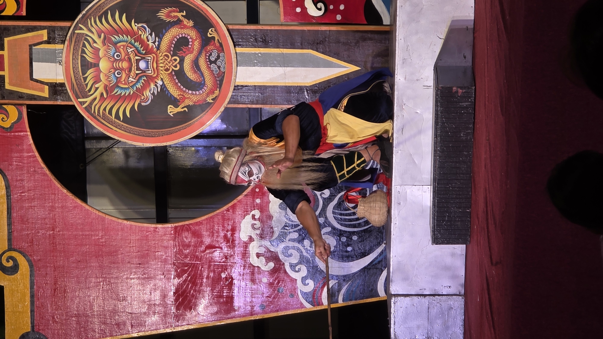The image captures a performance, possibly a theatrical production, occurring in what appears to be a venue in Nha Trang Ward, Vietnam. A central figure, dressed in traditional attire with a long white wig and painted face, is kneeling on a stage. The costume includes a black top, blue shoulder cloth, a yellow sash, and dark pants with yellow accents. He is holding a thin wooden stick. Behind him, a decorative backdrop features a large round panel with a dragon design and various painted patterns in red, white, and blue, including wave-like motifs. The stage includes both painted scenery and more utilitarian construction elements, with a section covered in dark red fabric suggesting a stage curtain.