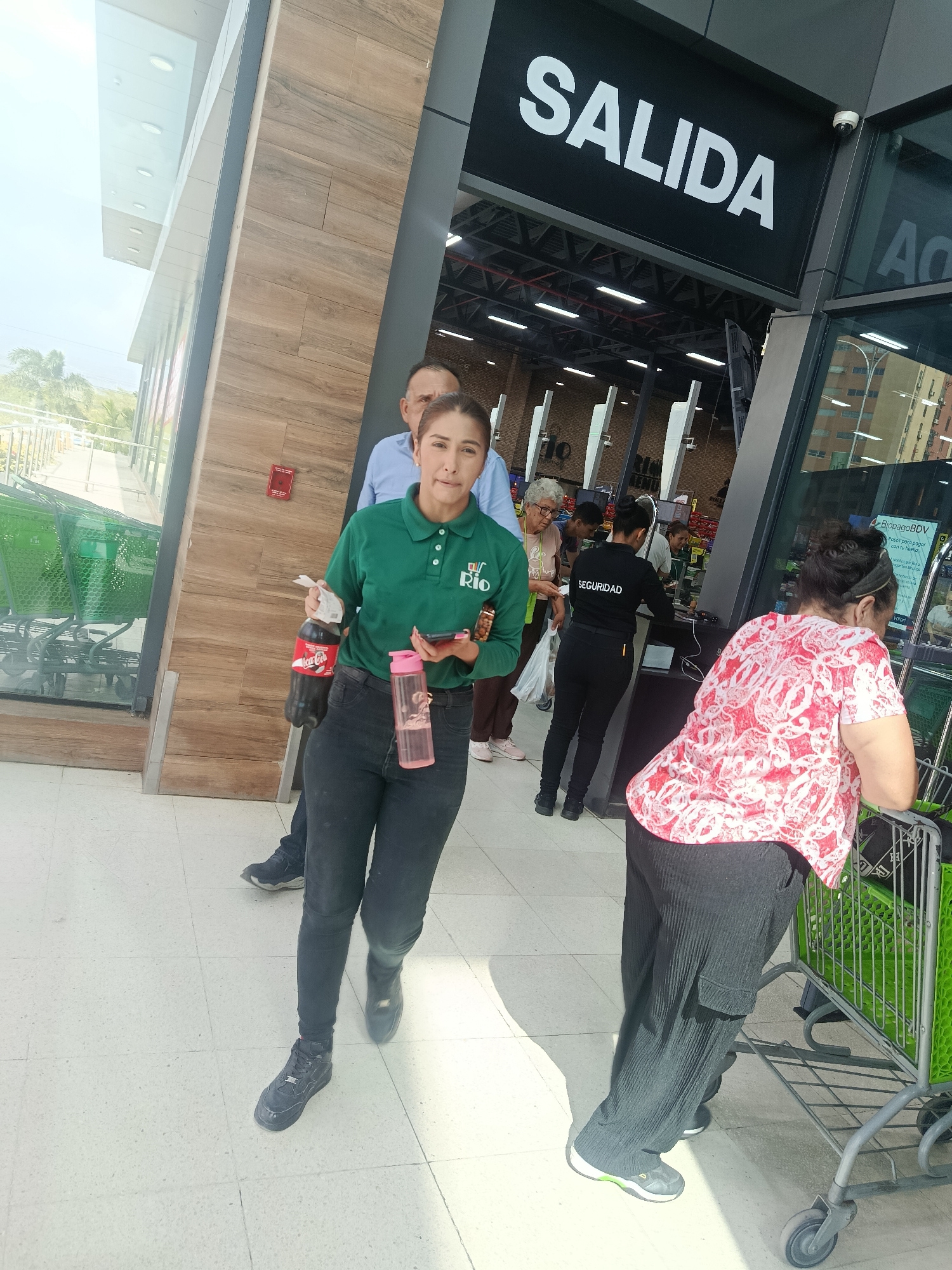 The image captures a bustling scene at the exit of a supermarket or large store, likely in Juan Griego, Venezuela, given the context of "BiopagoBDV." The time of day appears to be bright and sunny, indicating daytime.
In the foreground, a woman wearing a green polo shirt with a "Río" logo, black jeans, and black sneakers walks towards the viewer. She holds a bottle of Coca-Cola in one hand, a pink water bottle, and a smartphone in the other. Behind her, a man in a light blue shirt is partially visible. To the right, another woman in a red and white patterned shirt and dark, wide-leg pants leans on a bright green shopping cart.
Further inside the store, visible through the entrance/exit, a security guard in a black uniform with "SEGURIDAD" printed on the back faces away from the camera, attending to a customer at a counter. Other customers are also seen within the store, along with more green shopping carts.
Above the exit, a prominent black sign with white lettering clearly reads "SALIDA" (Exit). Another sign inside the store, partially visible, mentions "BiopagoBDV" and "Pasos para pagar con tu huella" (Steps to pay with your fingerprint), indicating a biometric payment system. The building's exterior features modern architecture with a wooden-textured facade and large glass panels. A red fire alarm is mounted on a pillar. The floor is a light-colored tiled surface.