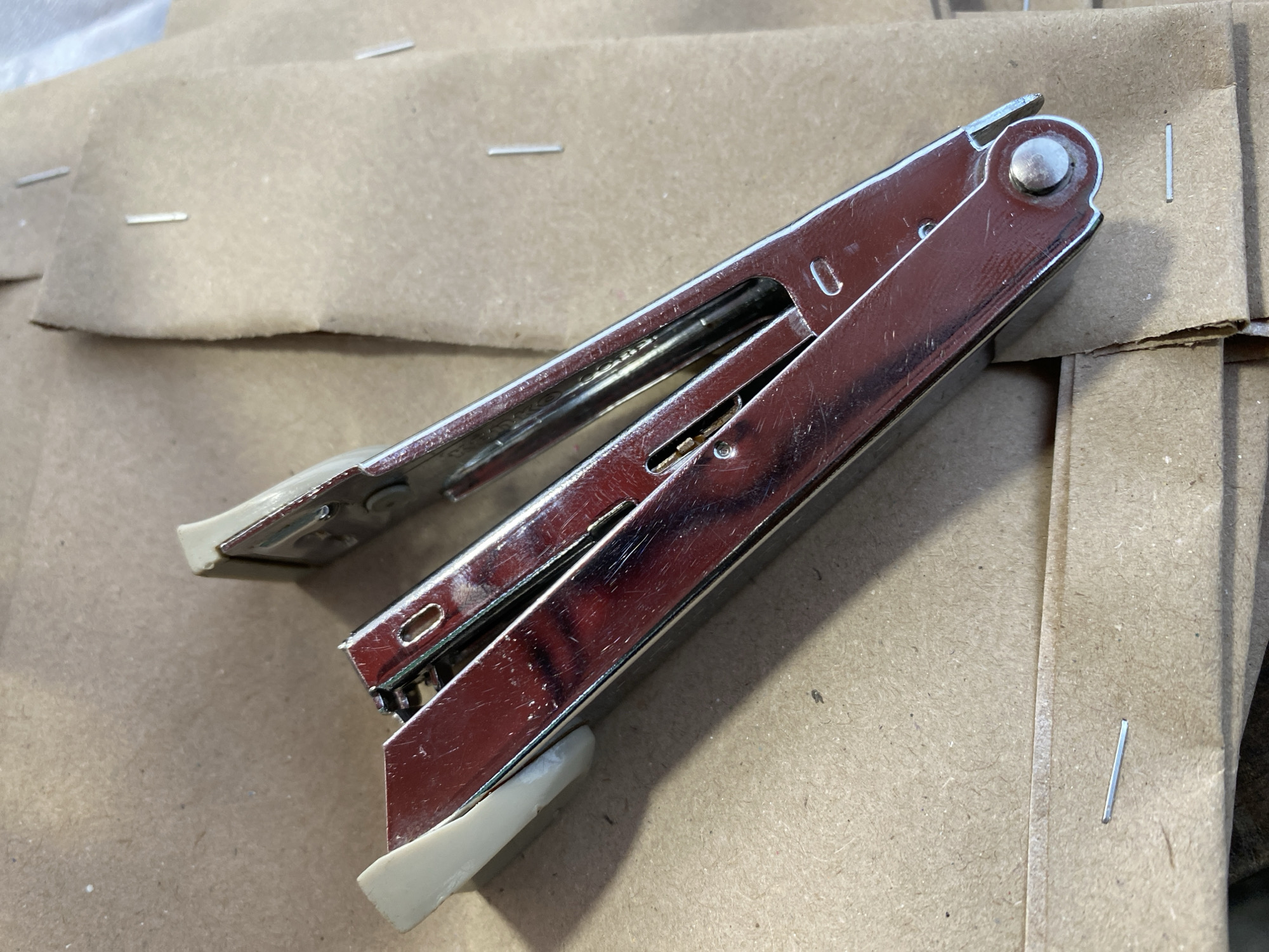 A close-up, overhead shot shows a shiny, chrome stapler with beige plastic accents on a light brown, cardboard surface. The stapler is open, revealing its inner workings. The cardboard surface has visible texture and appears to be part of a larger object or stack. Several individual staples are scattered on the cardboard, some clearly visible and others barely noticeable. The lighting is bright, creating a slight glare on the stapler's metal surface and casting subtle shadows. There is no indication of the city of Kasihan, Indonesia, or any other specific location in the image.
