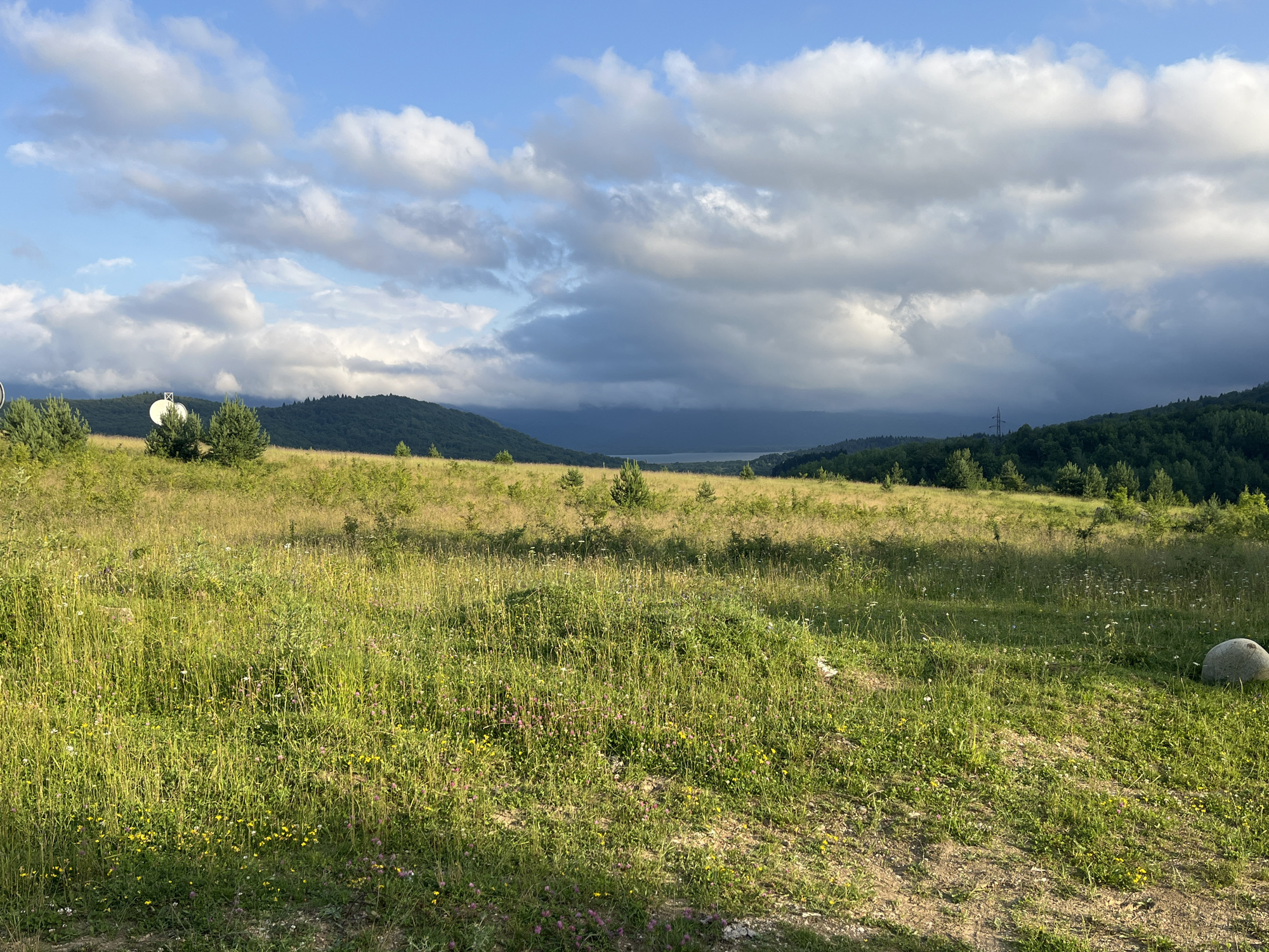 An outdoor, wide-angle shot captures a rolling landscape under a cloudy sky. In the foreground, a dirt path leads into a meadow filled with tall green grass and scattered wildflowers, including small yellow and pink blossoms. A large, grey boulder rests on the right side of the path. The middle ground features a more sparsely vegetated field with small evergreen trees and a prominent white satellite dish with a cross symbol. Beyond this, rolling green hills covered in dense forest ascend, with a body of water visible in the distance between two of the hills. The sky is a mix of blue and dramatic grey clouds, with sunlight breaking through in places, illuminating the landscape. The scene appears to be located near Nikortsminda, Georgia, based on the surrounding mountainous terrain and vegetation.