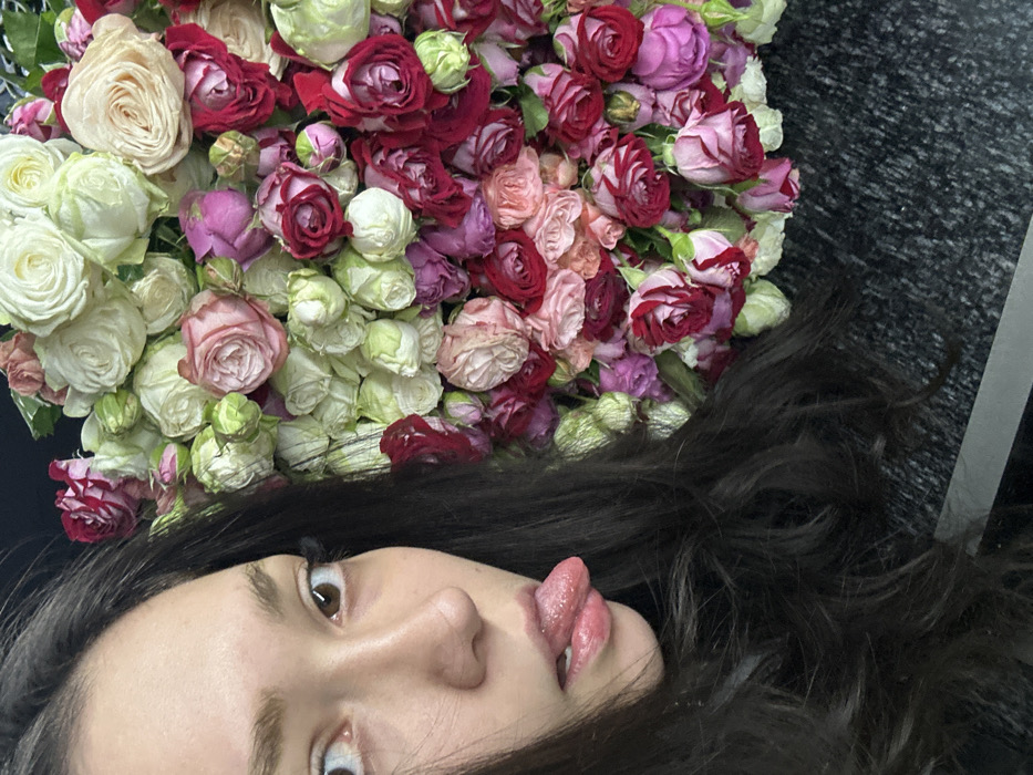 A woman lies on her side with dark hair cascading around her face. She sticks out her tongue, revealing a double split. An abundant bouquet of miniature roses in shades of white, pink, and deep red sits above her head. The blossoms are densely packed and appear fresh. The background is a dark, mottled surface, possibly fabric or carpet.