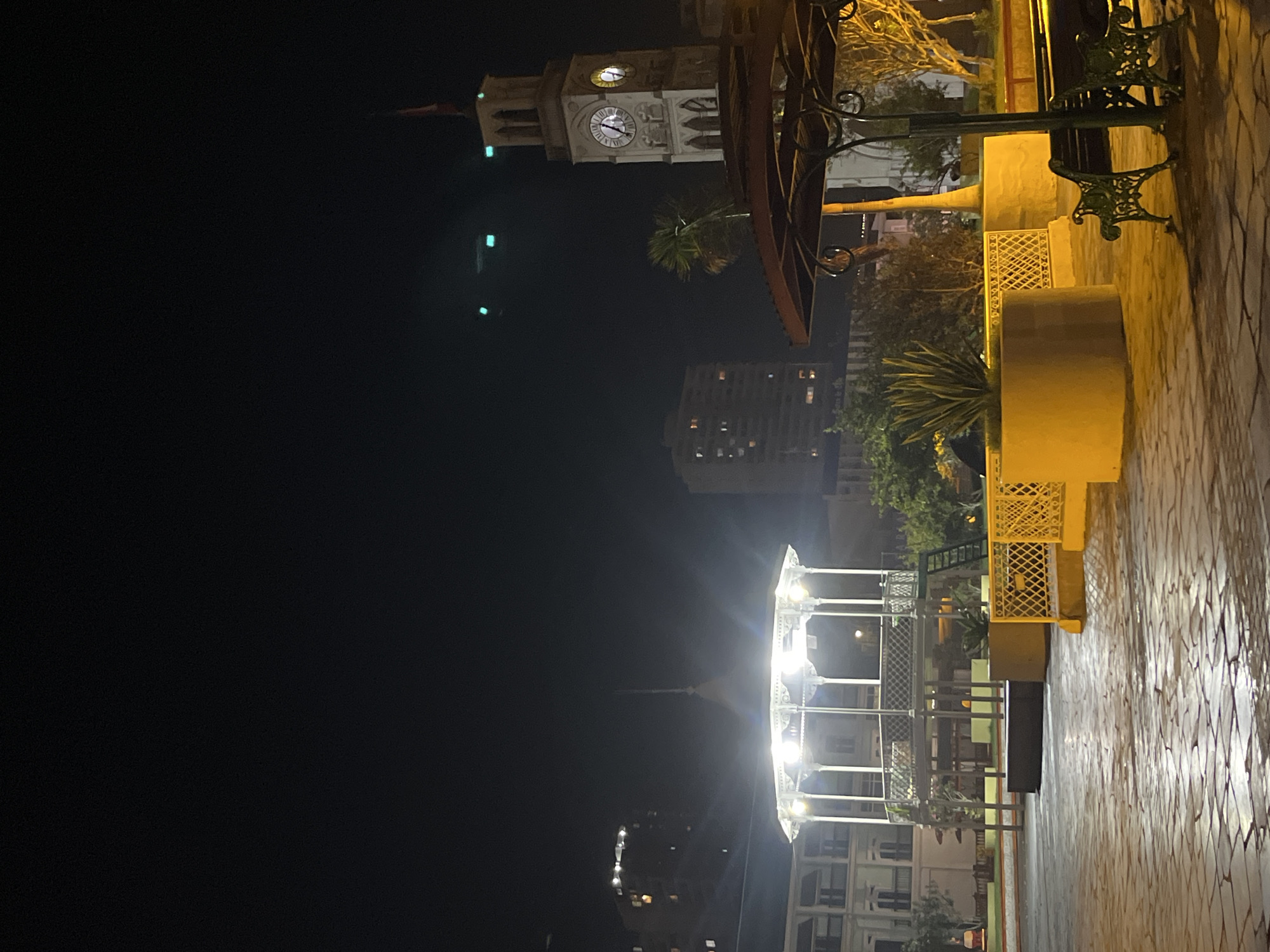 The image shows a cityscape at night, likely a public square or park. The ground is paved with stone tiles. In the background, there is a clock tower and a tall building with lighted windows. In the foreground, there is a gazebo illuminated with bright lights, and seating areas with ornate metal benches and planters. There are trees and other greenery visible around the square. The overall lighting is dim, with bright spots from the illuminated structures.