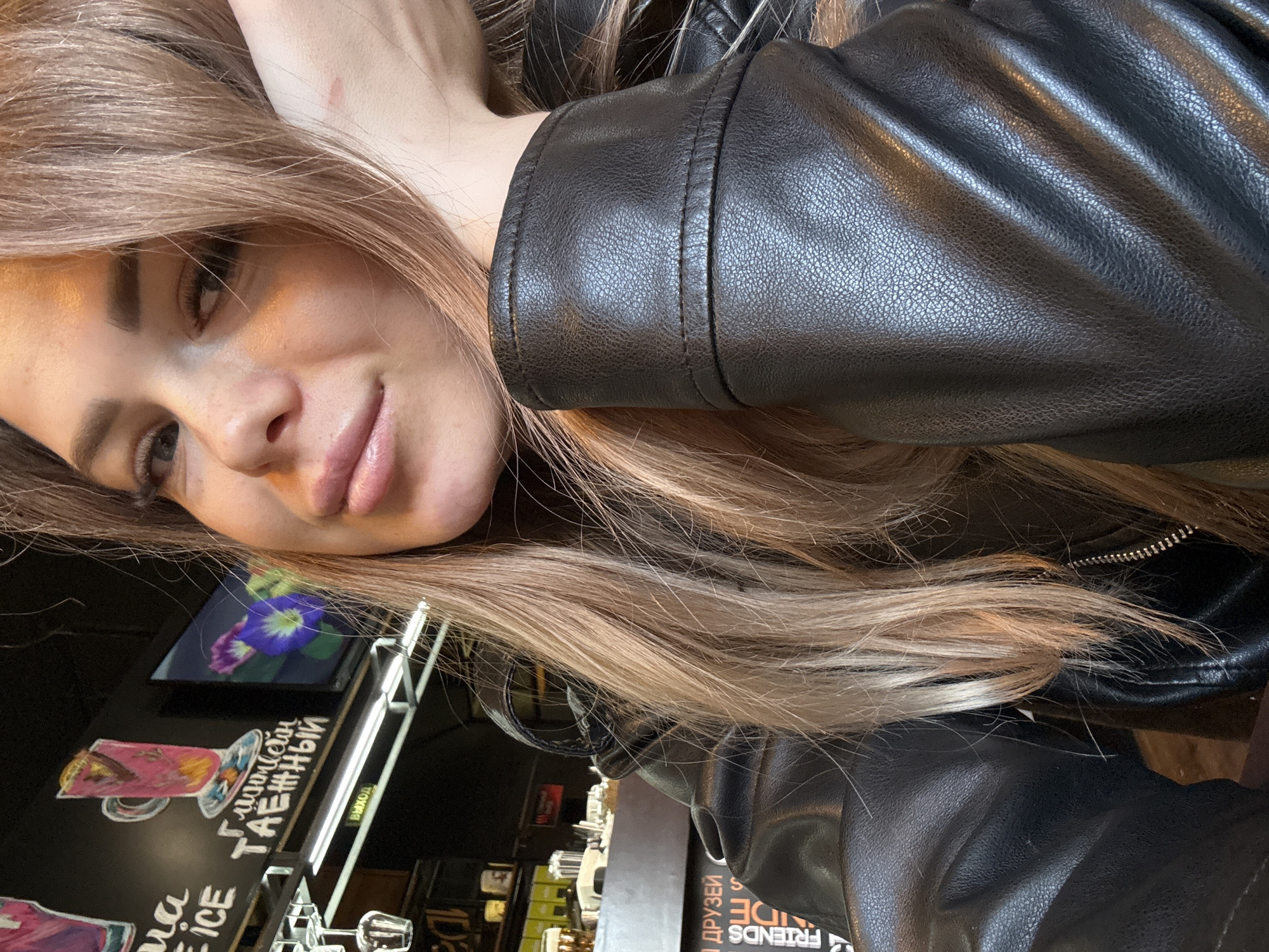A young woman with long, light brown hair, parted on the side, is captured in a close-up, rotated selfie. She is lying down, likely on her left side, with her head resting on her left arm. She looks directly at the camera with a slight, relaxed smile. Her face shows natural make-up, defined eyebrows, and long eyelashes. A small, light red mark is visible on her upper arm or shoulder, partially covered by her hair.
She is wearing a dark, presumably black, leather-textured jacket. The material appears supple with fine grain and visible stitching details on the sleeves. A metallic zipper is also visible on the jacket.
The background suggests an indoor setting, likely a casual restaurant, cafe, or bar, indicated by dark surfaces, displays, and metallic fixtures. On the left side of the frame, there's a dark chalkboard-like surface with colorful drawings and white text. One drawing depicts a pink cocktail in a glass. Visible text includes "ГЛИКАЙ ТАЁЖНЫЙ" (GLIKAY TAYOZHNYY, likely a drink name) and partially visible "ICE". A small, illuminated sign displays "ВЫХОД" (VYKHOD), meaning "EXIT". Towards the bottom right, reversed text can be seen, including "ДРУЗЕЙ" (DRUZEY, meaning "FRIENDS") and "SIDE", possibly part of a sign like "FRIENDS' SIDE". The lighting appears to be ambient indoor light, casting some highlights on the jacket. The overall mood is casual and relaxed.