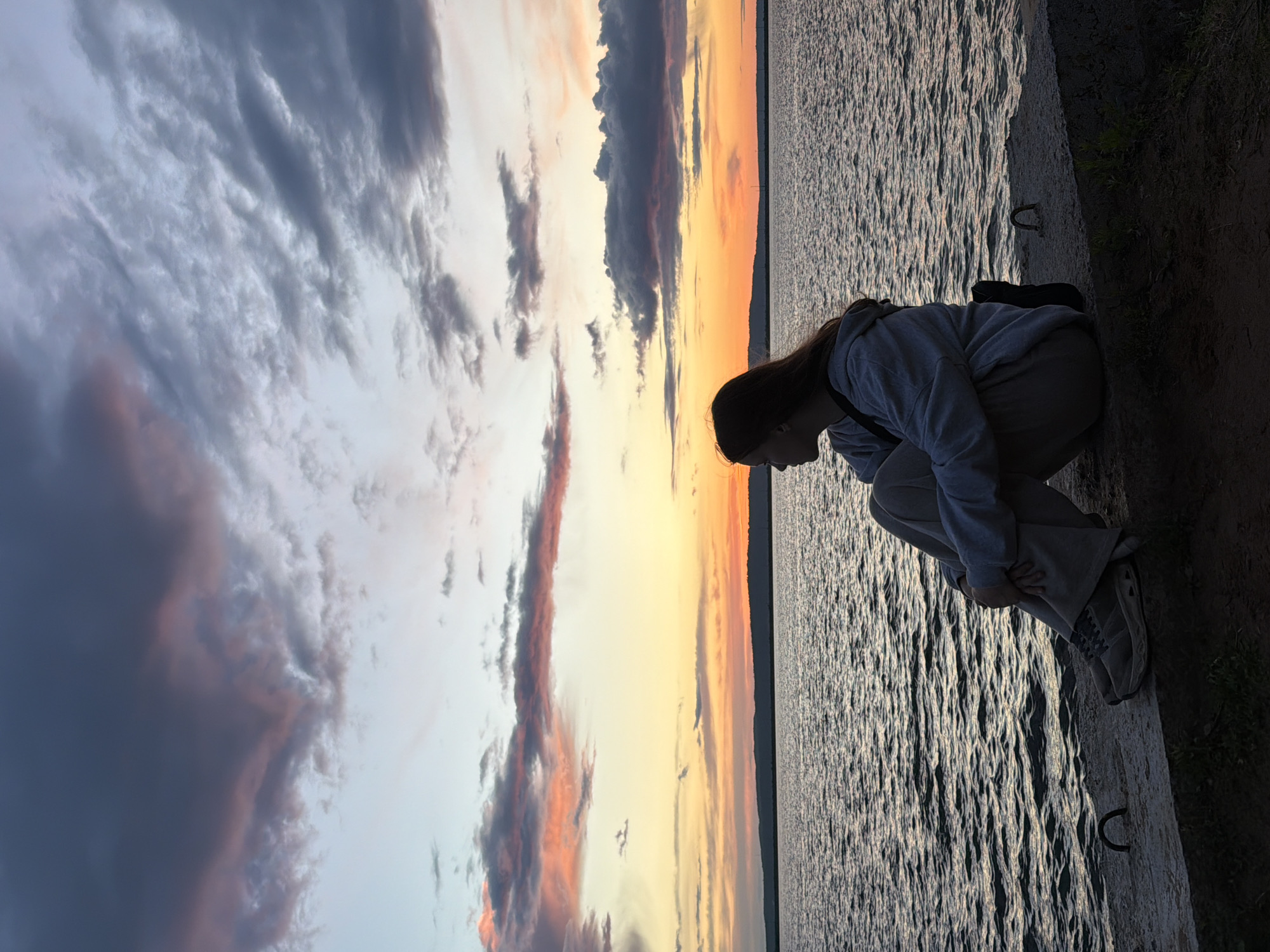 A person is crouching at the edge of a body of water, likely the Kama River, at Chaikovsky, Russia, evidenced by the river's distinctive characteristics and the setting sun alignment. The individual is wearing a light-colored hooded sweatshirt, trousers, and sneakers. They are positioned near what appears to be a concrete embankment or walkway. The sky features dramatic cloud formations with colors indicating a sunset or sunrise. The water reflects the light, creating a textured surface.