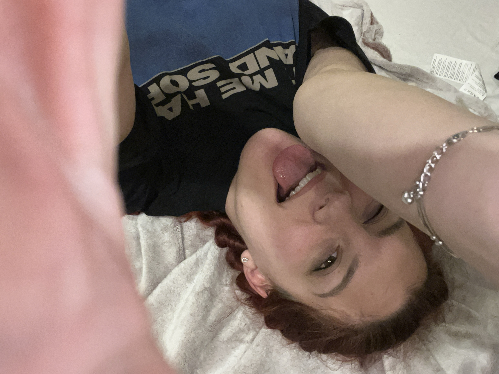 A woman with reddish-brown hair is lying on a white textured blanket. She is wearing a black T-shirt with blue lettering. Her tongue is sticking out, and she is looking towards the viewer. A silver bracelet is on her right arm, and she has earrings in her ears. The image appears to be taken indoors. The location is not discernible from the image, so it's not possible to confirm the city of Sofia, Bulgaria.