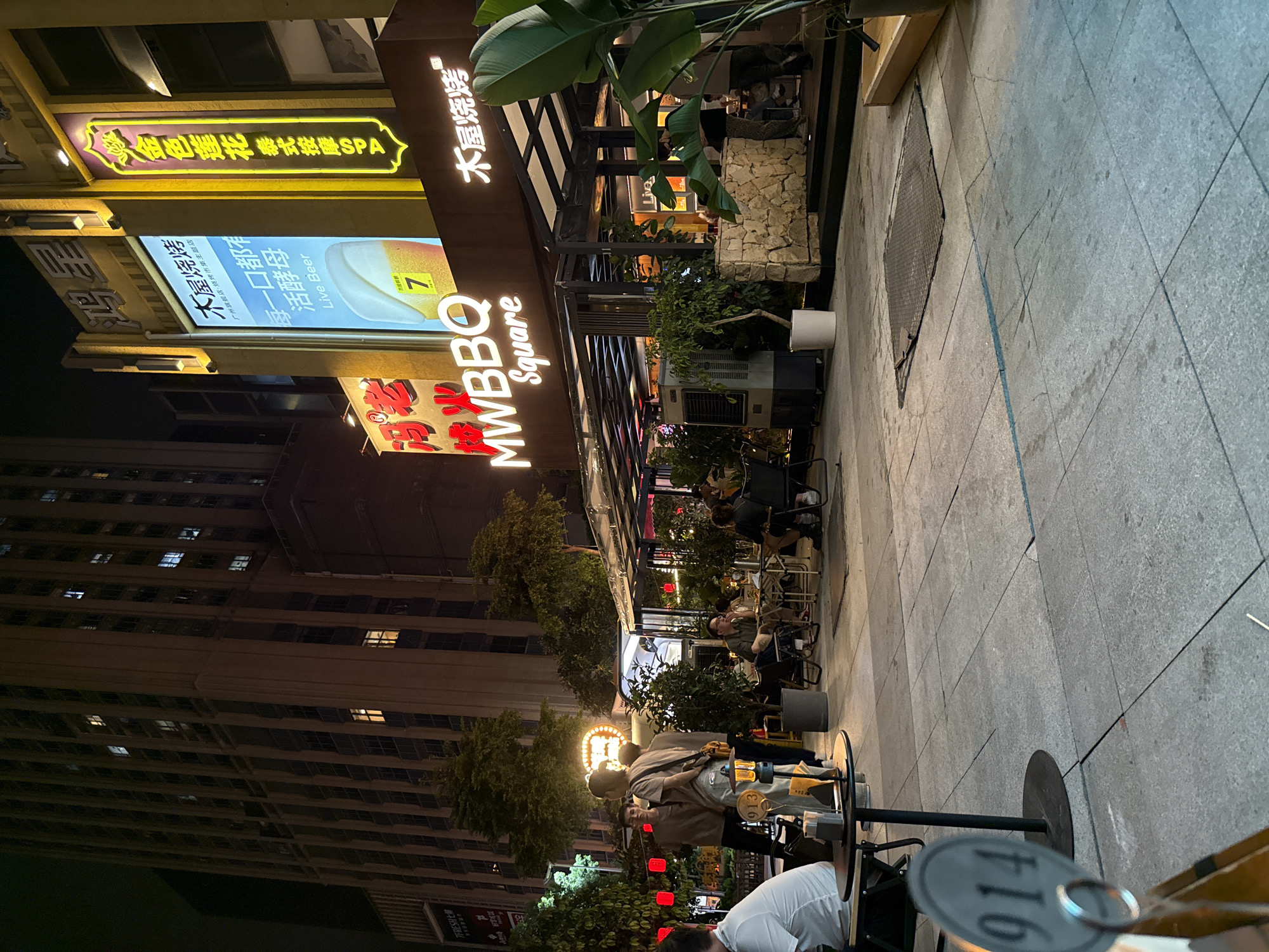 The image depicts a nighttime street scene in Haizhu District, China, featuring an outdoor dining area for a restaurant called "MWBBQ Square." Several people are seated at tables under a covered patio, engaged in dining and conversation. The restaurant's illuminated sign, "MWBBQ Square," is prominently displayed. Adjacent to the restaurant, a tall building with numerous lit windows looms in the background, suggesting an urban environment. Further signage in Chinese characters, including one for "Golden Lotus Thai Massage SPA," indicates the presence of other businesses nearby. Red lanterns are visible hanging from trees, adding to the ambient lighting. The ground is paved with large stone tiles. A numbered table marker, "914," is visible in the foreground on a small round table. The overall scene conveys a bustling atmosphere of a commercial and entertainment district at night.