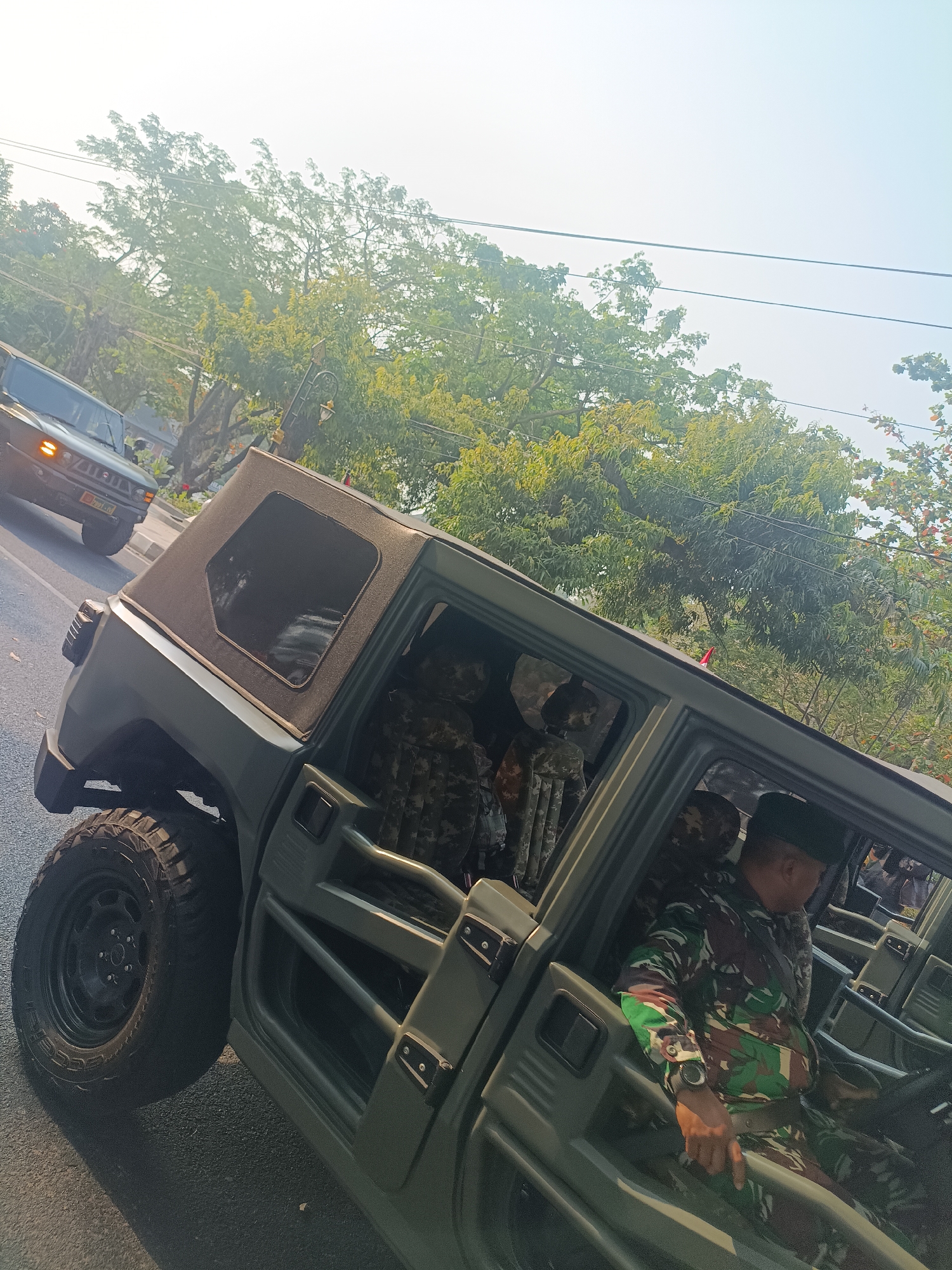 A man in camouflage uniform is sitting in the driver's seat of an olive-drab military vehicle, possibly a Humvee, in the city of Sleman, Indonesia. He is wearing a black cap and a watch on his left wrist. The vehicle has a soft top and visible roll bars. In the background, another similar military vehicle is visible driving on the road, and lush green trees line the street. The sky is bright and hazy, suggesting it is daytime. The scene appears to be a street or road in an urban or suburban environment.