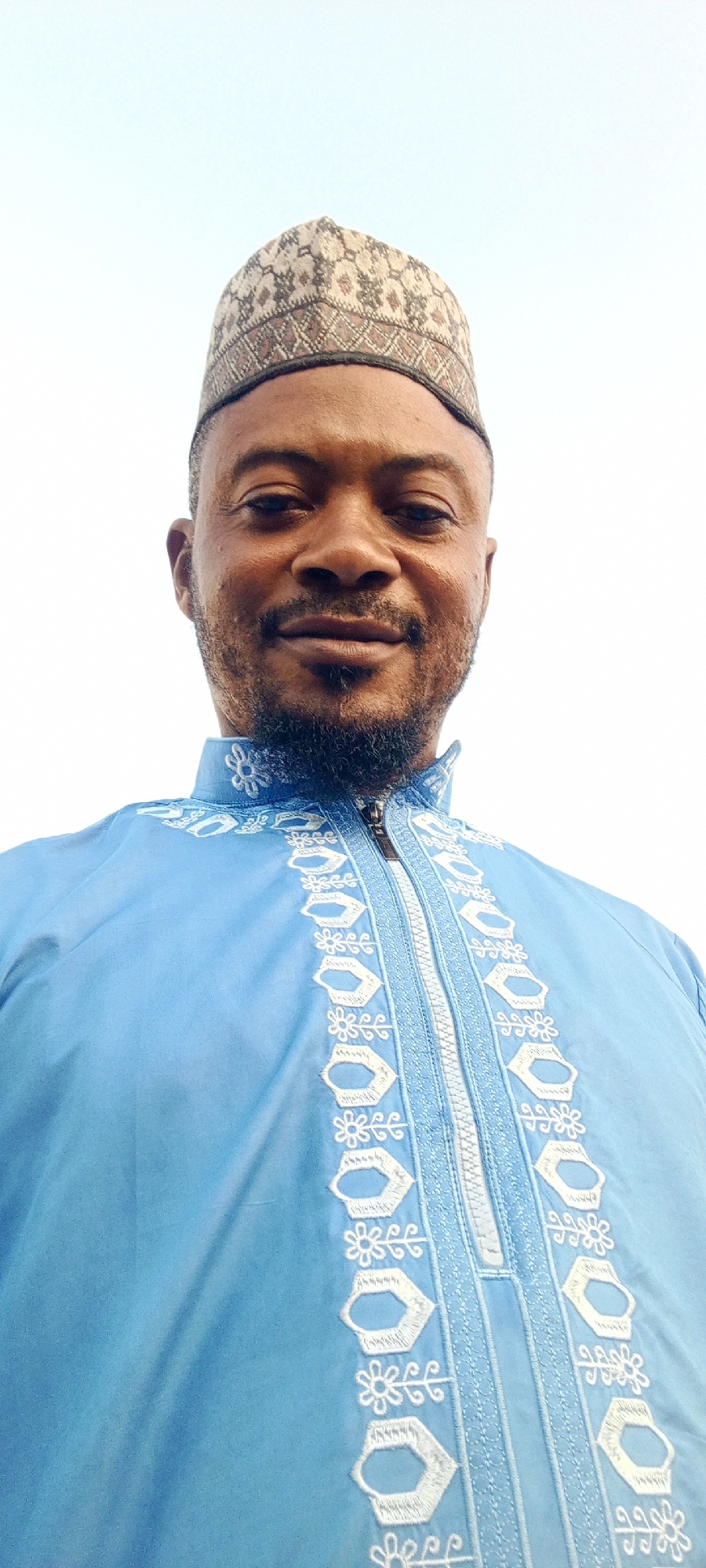 A close-up, low-angle portrait depicts an adult Black male subject against a plain, bright white to very light blue sky. The man has a short beard and mustache, dark eyes, and a neutral expression with a faint smile. He is wearing a light blue traditional garment, likely a tunic, featuring intricate white embroidery in floral and geometric patterns along the central placket and collar. The garment has a visible zipper closure at the neckline. On his head, he wears a multi-toned skullcap (kufi) with an intricate, repeating geometric pattern in shades of brown, beige, and black. The subject looks directly at the camera. The scene is outdoors in Gwagwarwa, Nigeria, under clear daylight conditions.