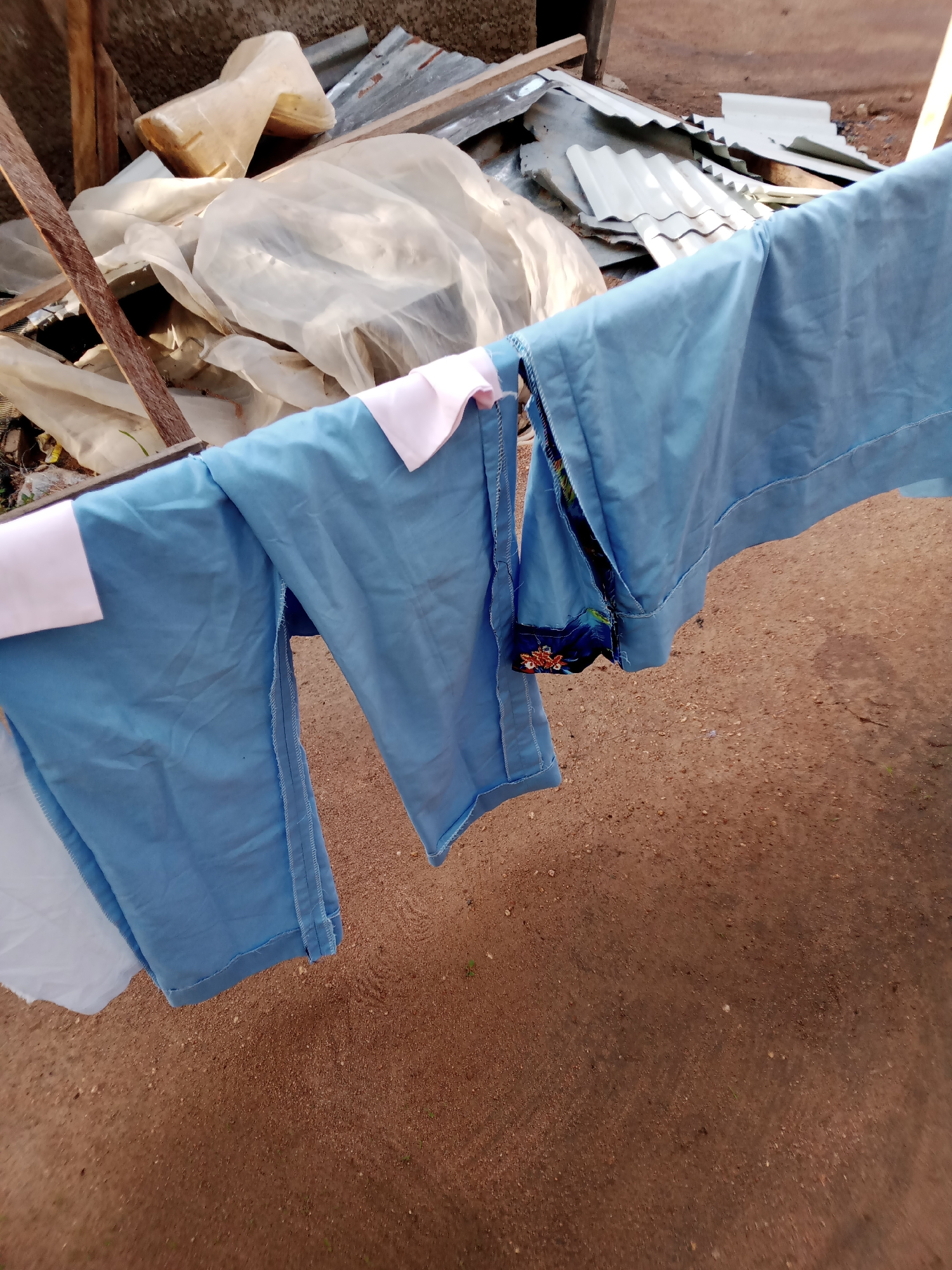 In Jauro Yinu, Nigeria, this image captures a close-up view of light blue garments, possibly uniforms, hanging on a clothesline. The fabric appears to be draped, with visible seams and hems. Some of the garments have white or pink accents, and one piece shows a dark fabric with a colorful pattern. In the background, a pile of white plastic sheeting and corrugated metal sheets are visible, suggesting an outdoor area that may be undergoing construction or storing materials. The ground is composed of reddish-brown dirt.